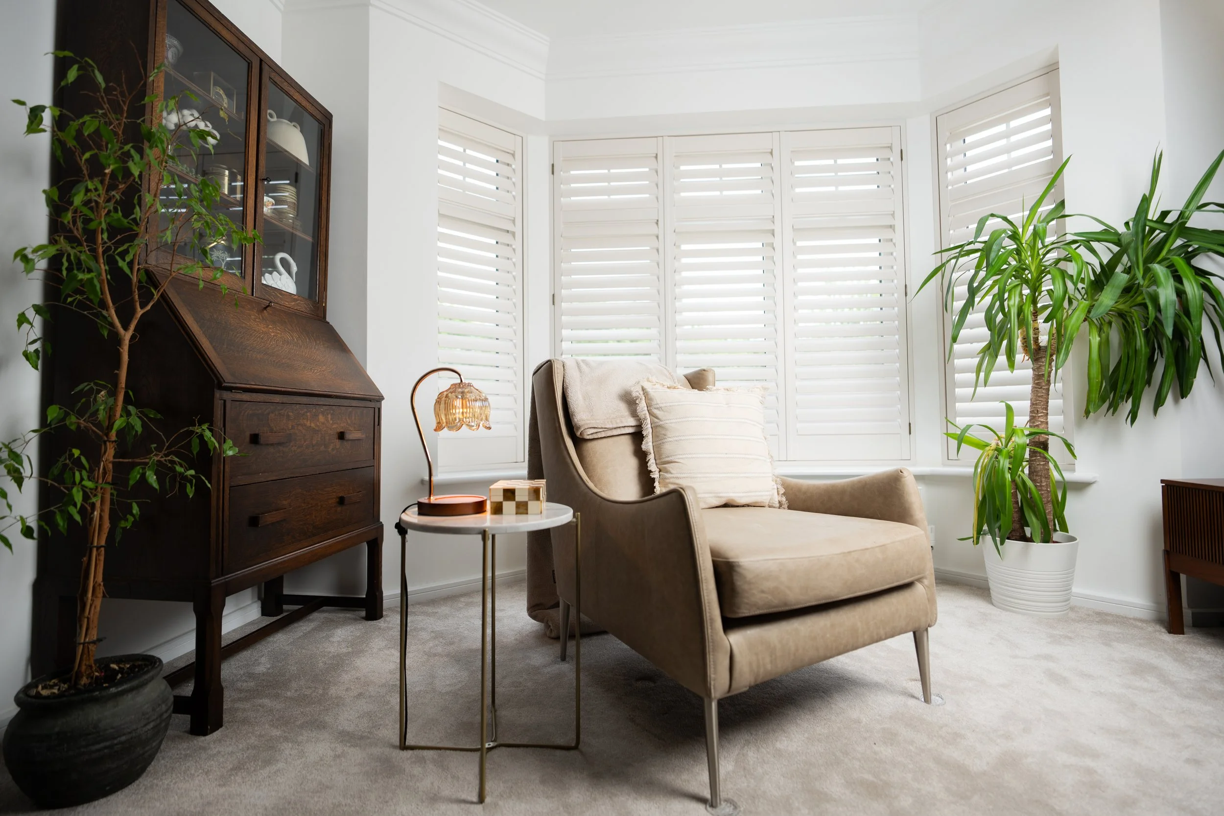 A cozy living room with a beige armchair, a small white side table with decorative items, a tall potted plant, a vintage wooden cabinet with glass doors displaying dishes, a floor lamp with a patterned shade, and large windows with white shutters let