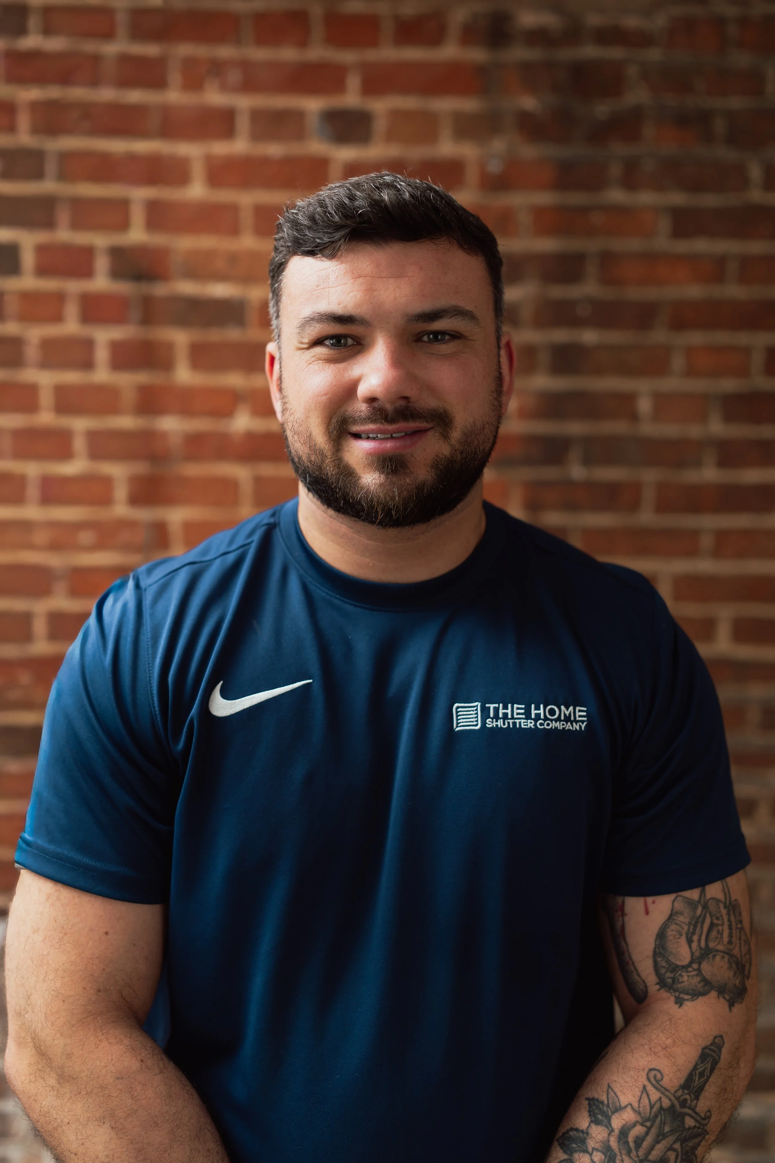 A man with dark hair and a beard is smiling at the camera, wearing a blue T-shirt with a logo and text, standing in front of a brick wall.