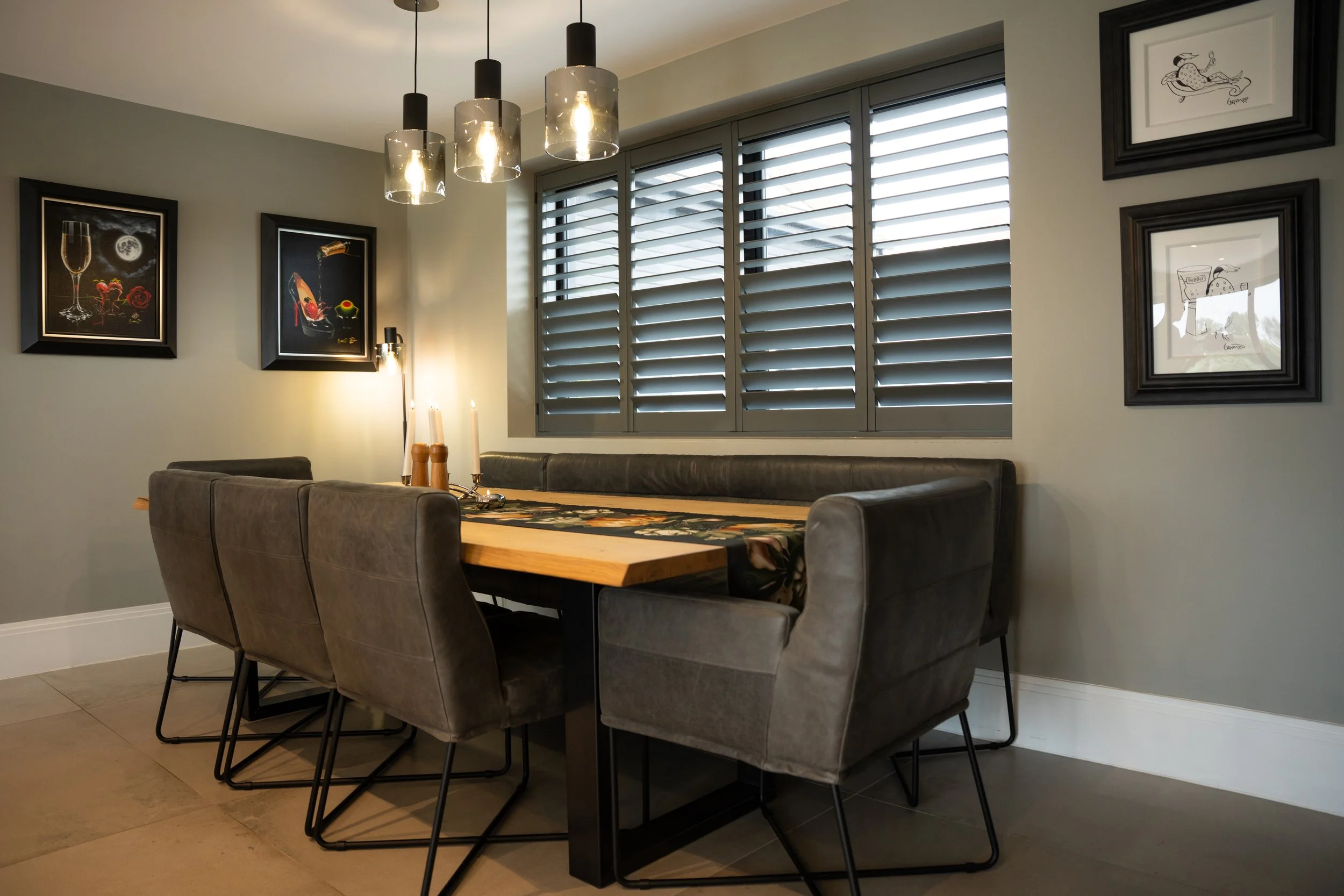 Dining room with a wooden table, six gray upholstered chairs, wall art depicting champagne and wine, a window with gray shutters, and modern pendant lighting.