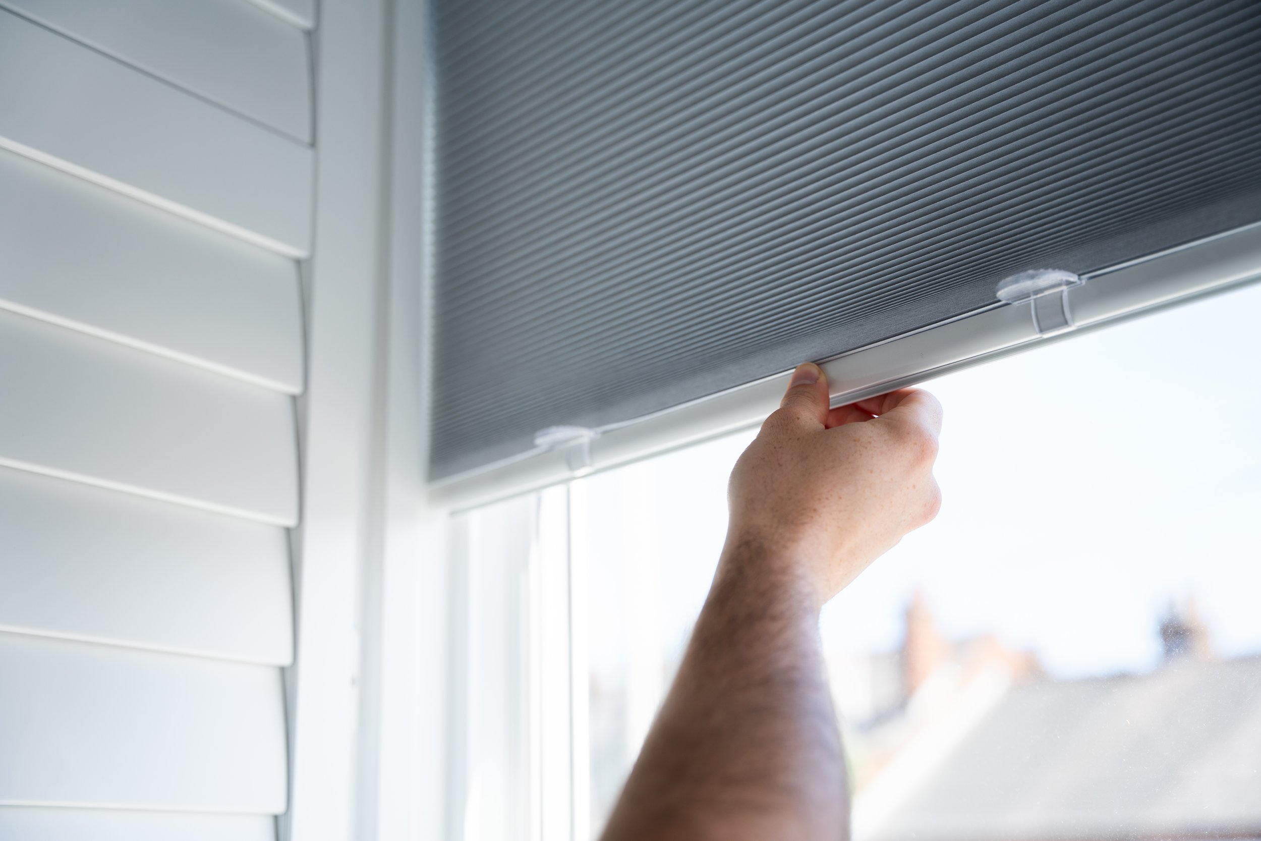 Close-up of a hand lifting an integrated window shutter blind on a white house exterior.