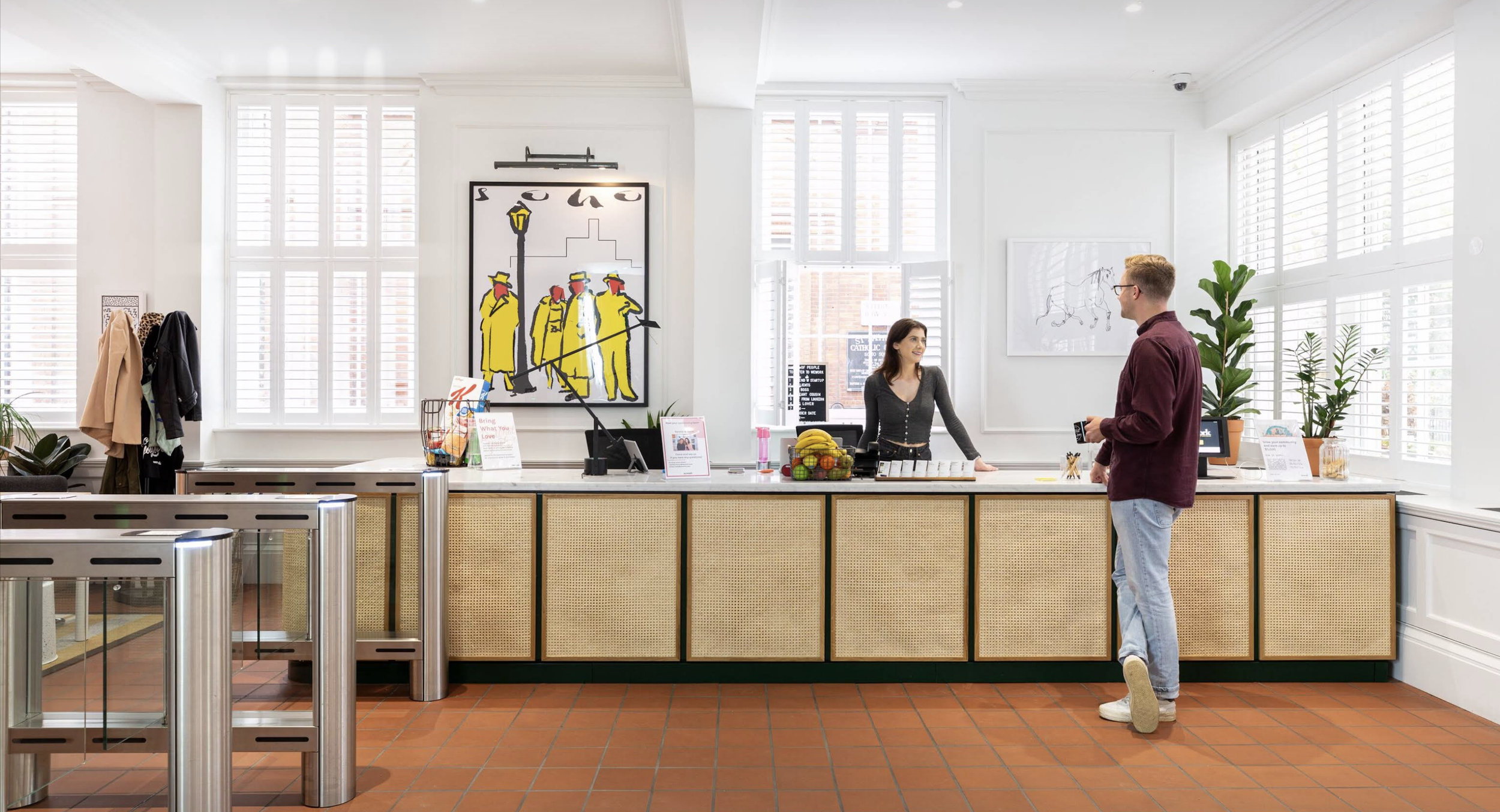 A modern hotel reception area with a man and woman engaging in conversation. The woman is behind the counter, smiling, with a basket of bananas and other items, while the man stands in front of her holding a key card. The space is bright with large w