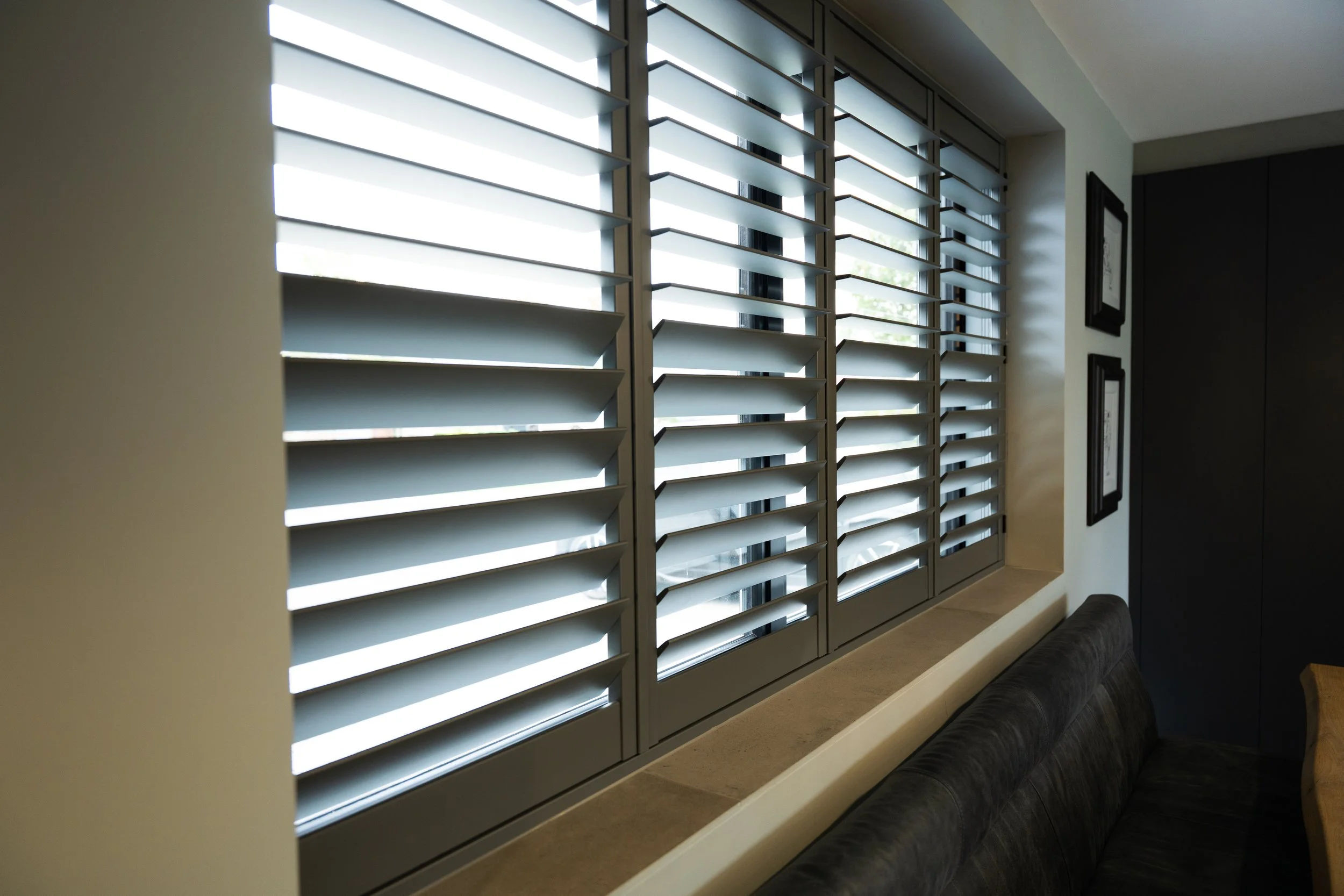 A window with dark grey shutters partially open, allowing light to enter. To the right, there are framed pictures on the wall, and a black padded bench or cushion placed along the wall beneath the window.