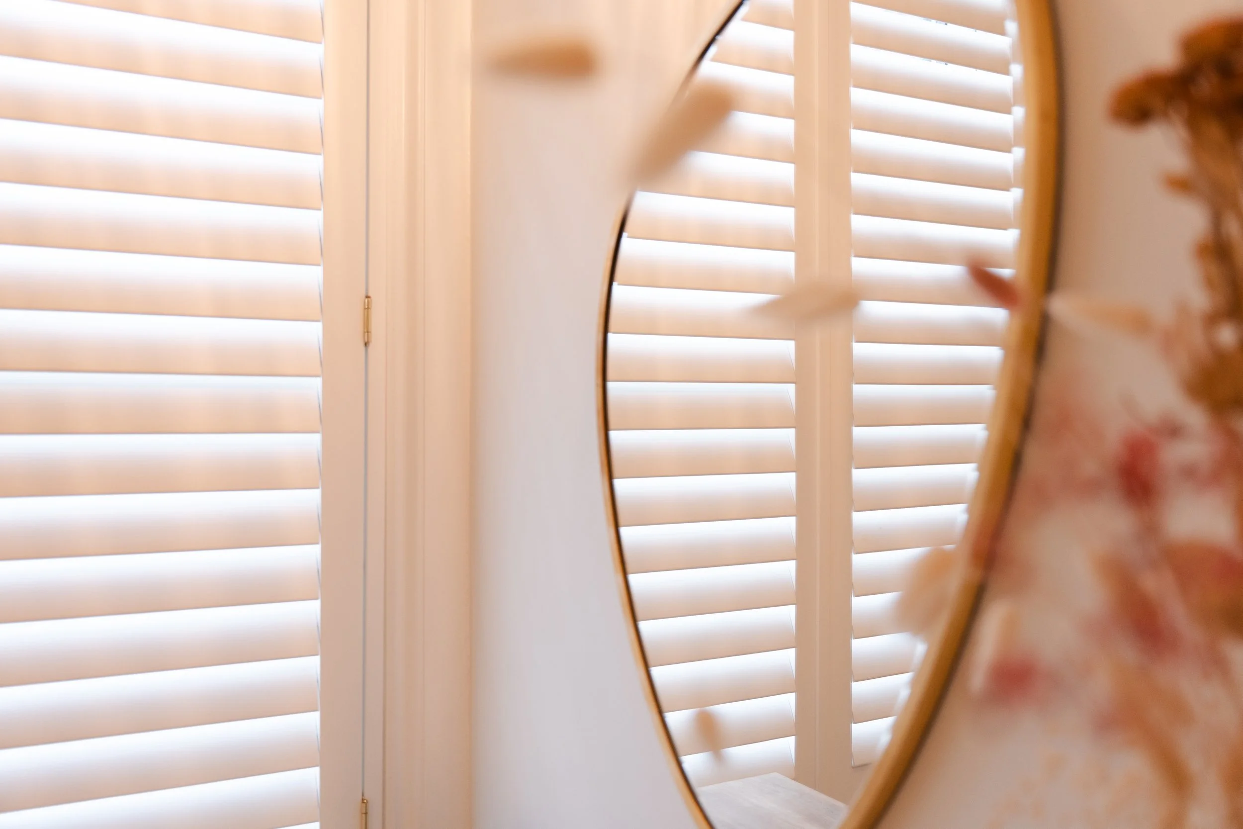 A mirror on a wall reflecting white wooden plantation shutters on a window.