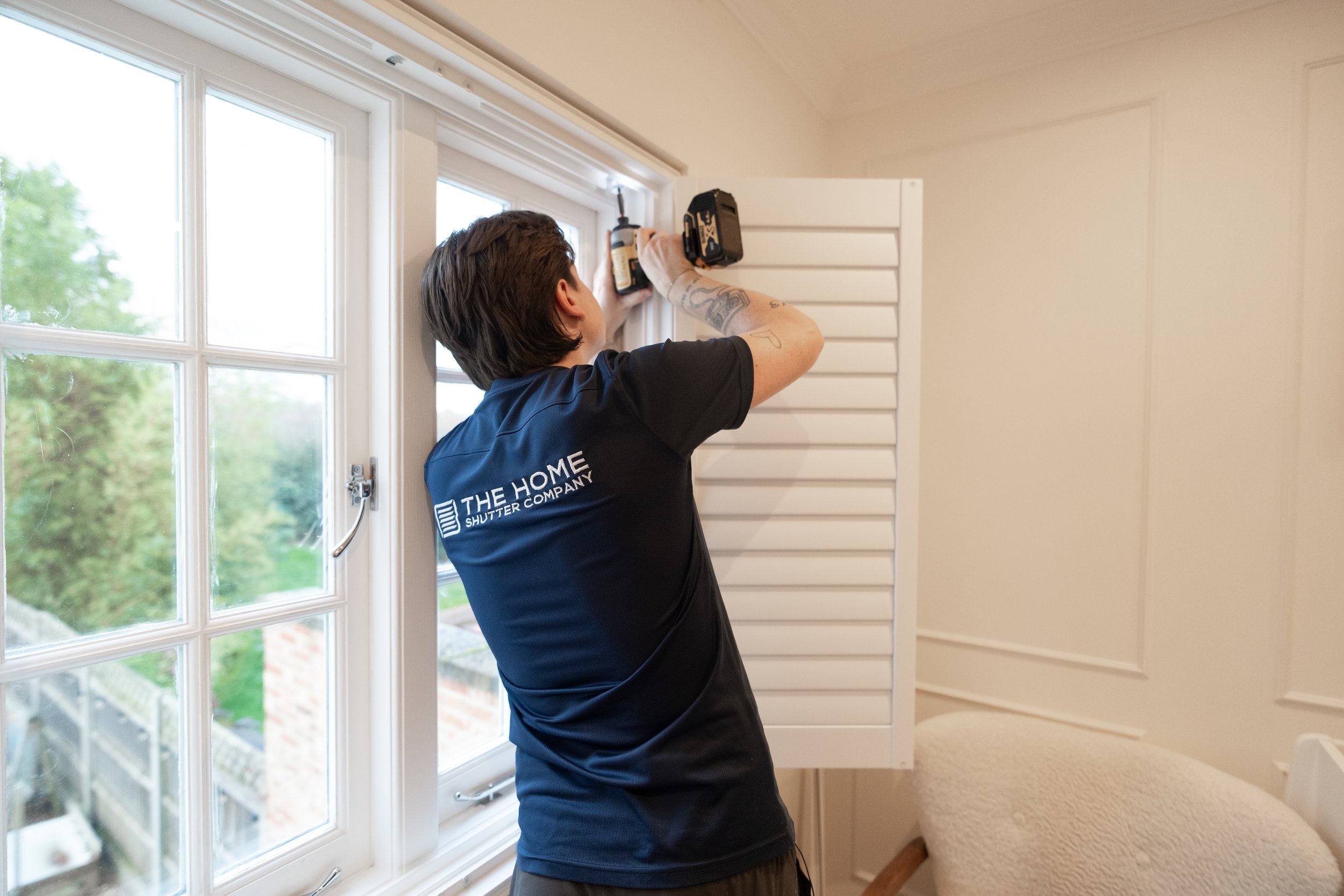 Person installing or repairing a window using a cordless drill, in a room with white walls and a large window.