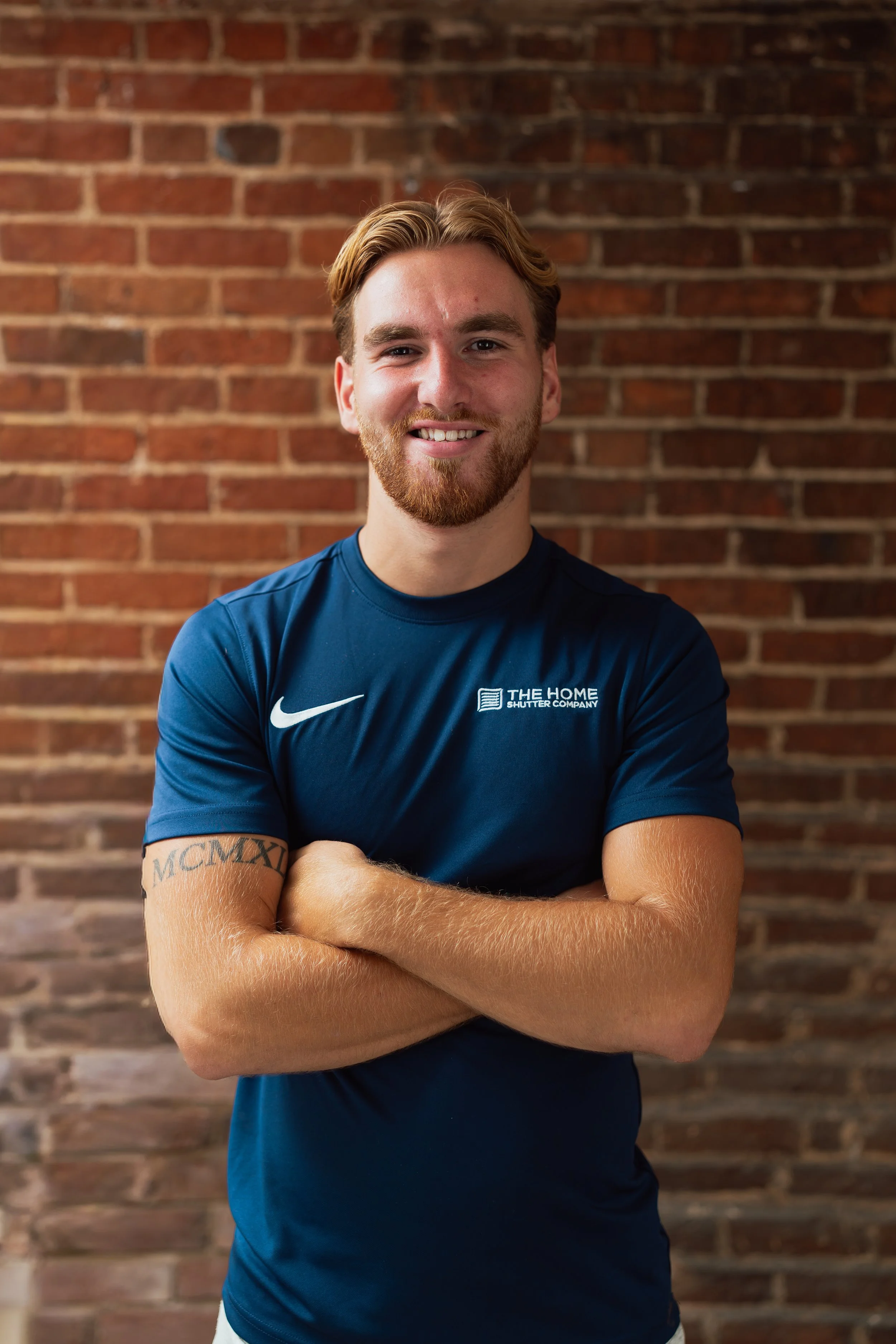 Portrait of a smiling young man with a beard and tattoos, standing with arms crossed in front of a brick wall, wearing a blue Nike athletic shirt.