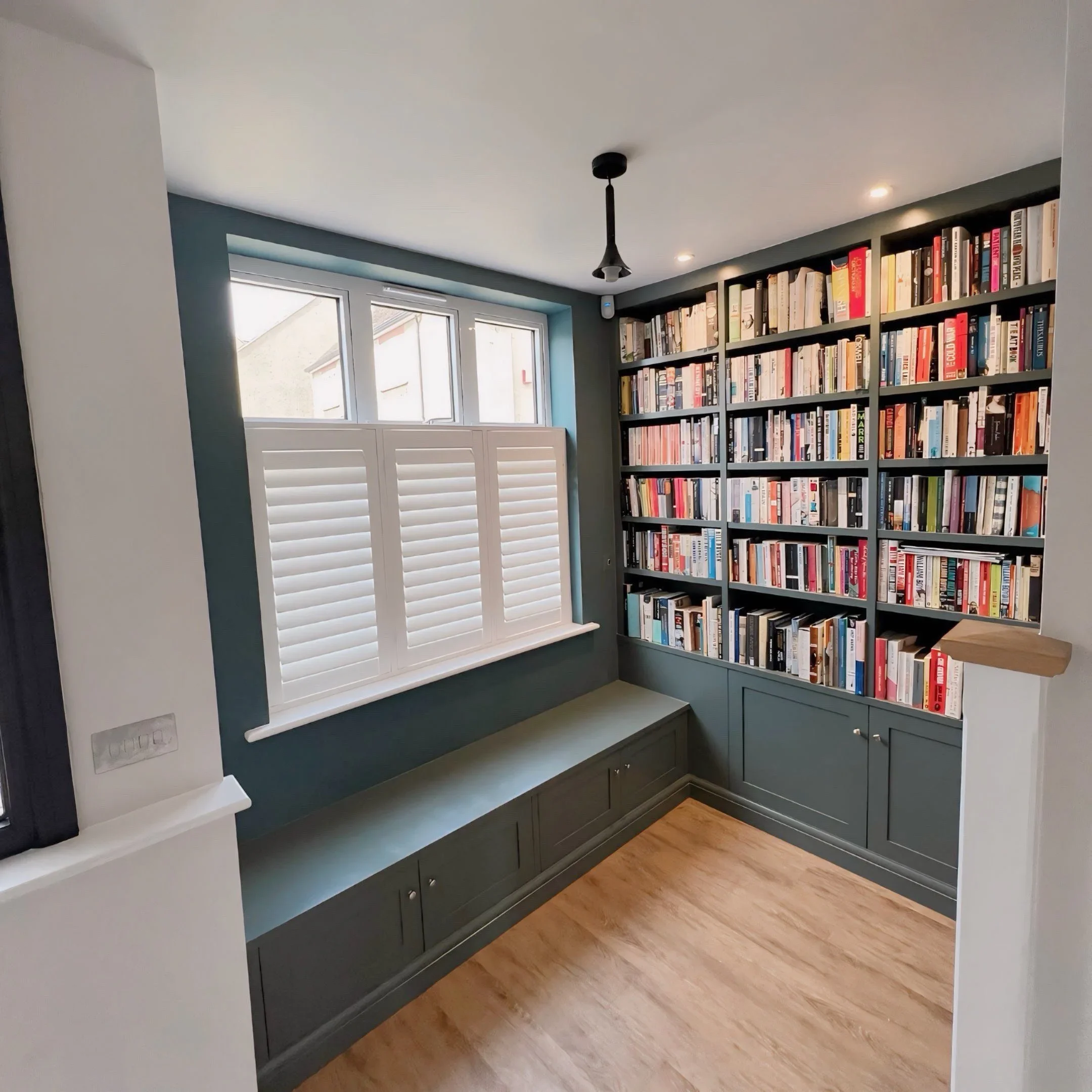 A room with a bookshelf filled with numerous books, a window with white shutters, a pendant ceiling light, green walls, and hardwood flooring.