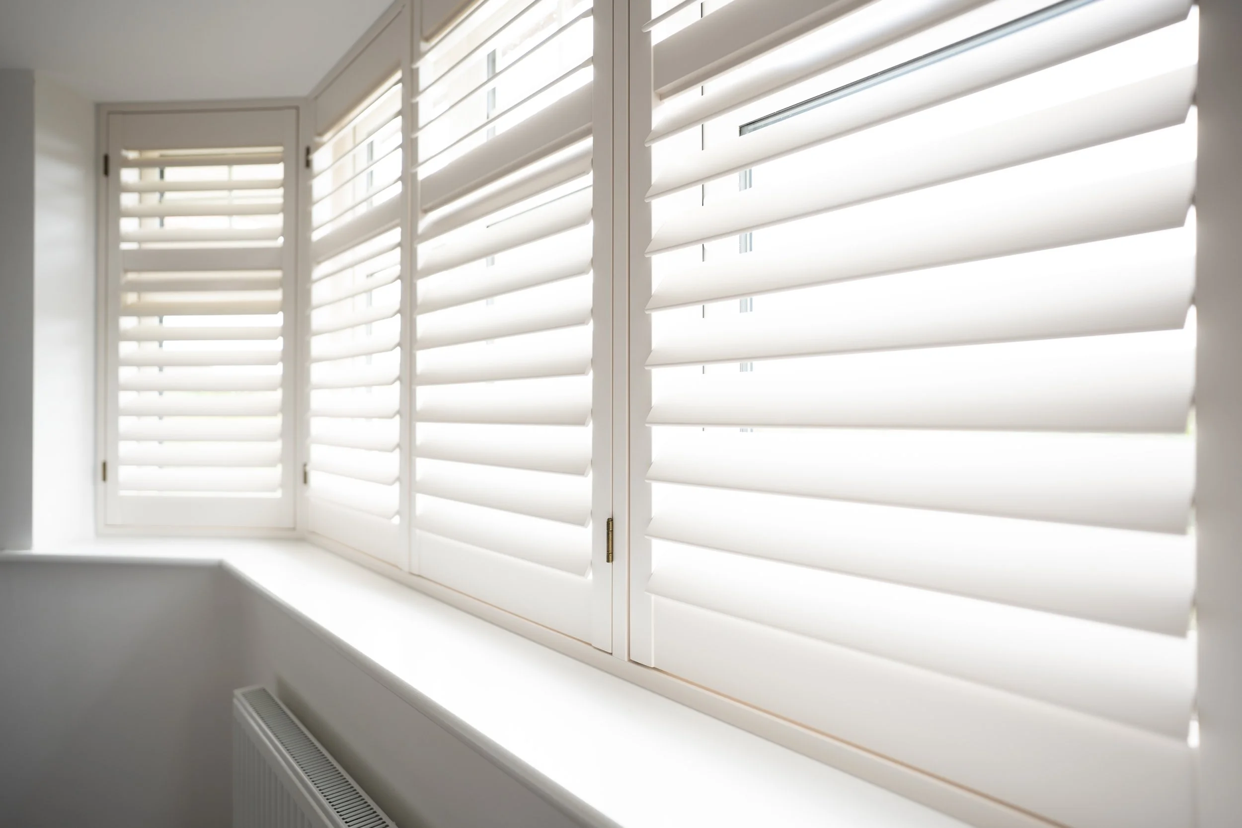 White plantation shutters on a window with sunlight shining through, inside a room.