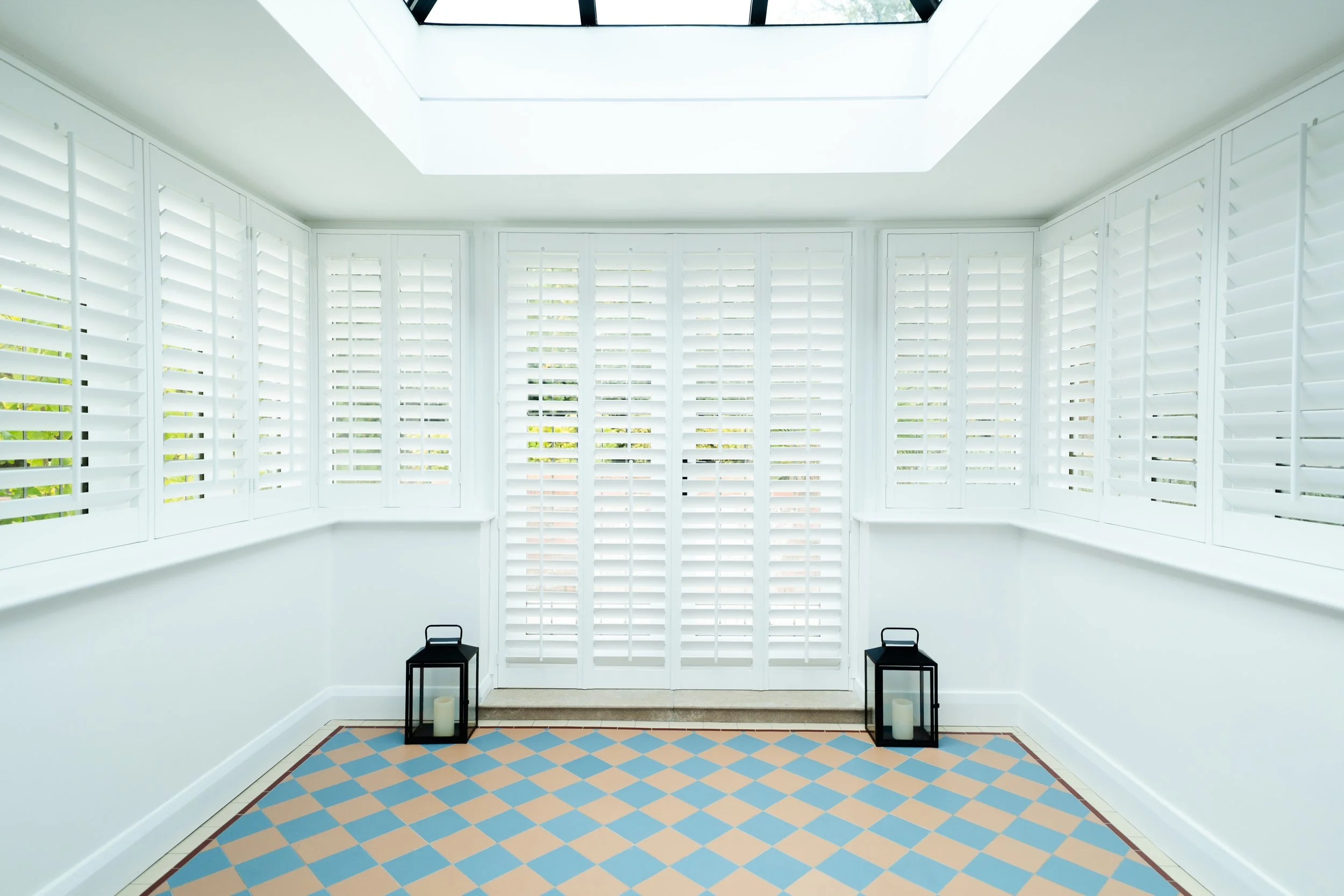 A bright white room with tall white plantation shutters on the windows, a skylight on the ceiling, and a patterned tile floor in shades of blue and beige. There are two black lantern-style candle holders with white candles on the floor beneath the wi
