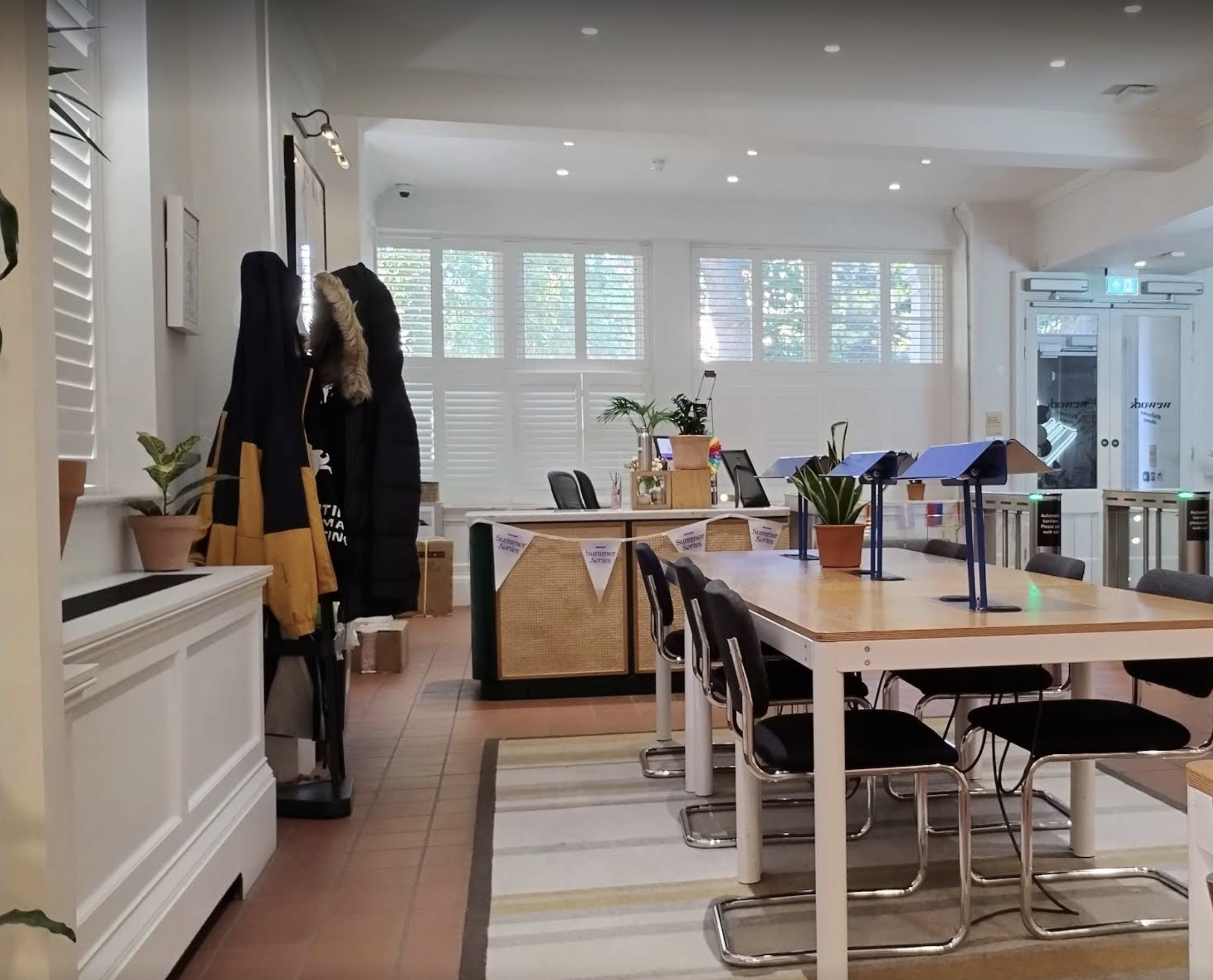 Interior of a modern, well-lit cafe or communal space with tables and chairs, including potted plants, coat hooks with jackets, and large windows with white shutters.
