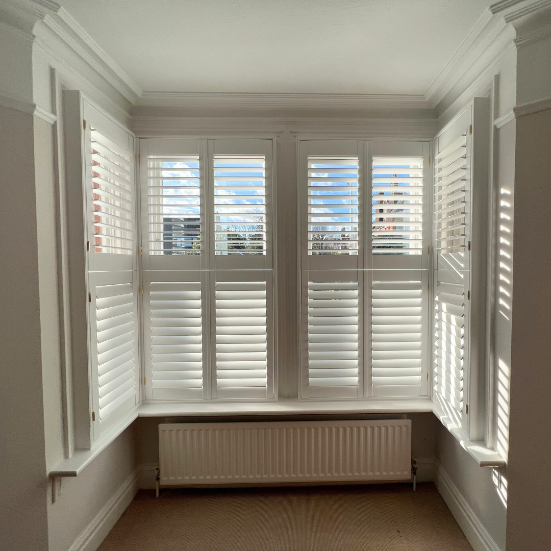 A cozy room with white plantation shutters covering large windows, a white radiator below the windows, white walls, and a beige carpeted floor.