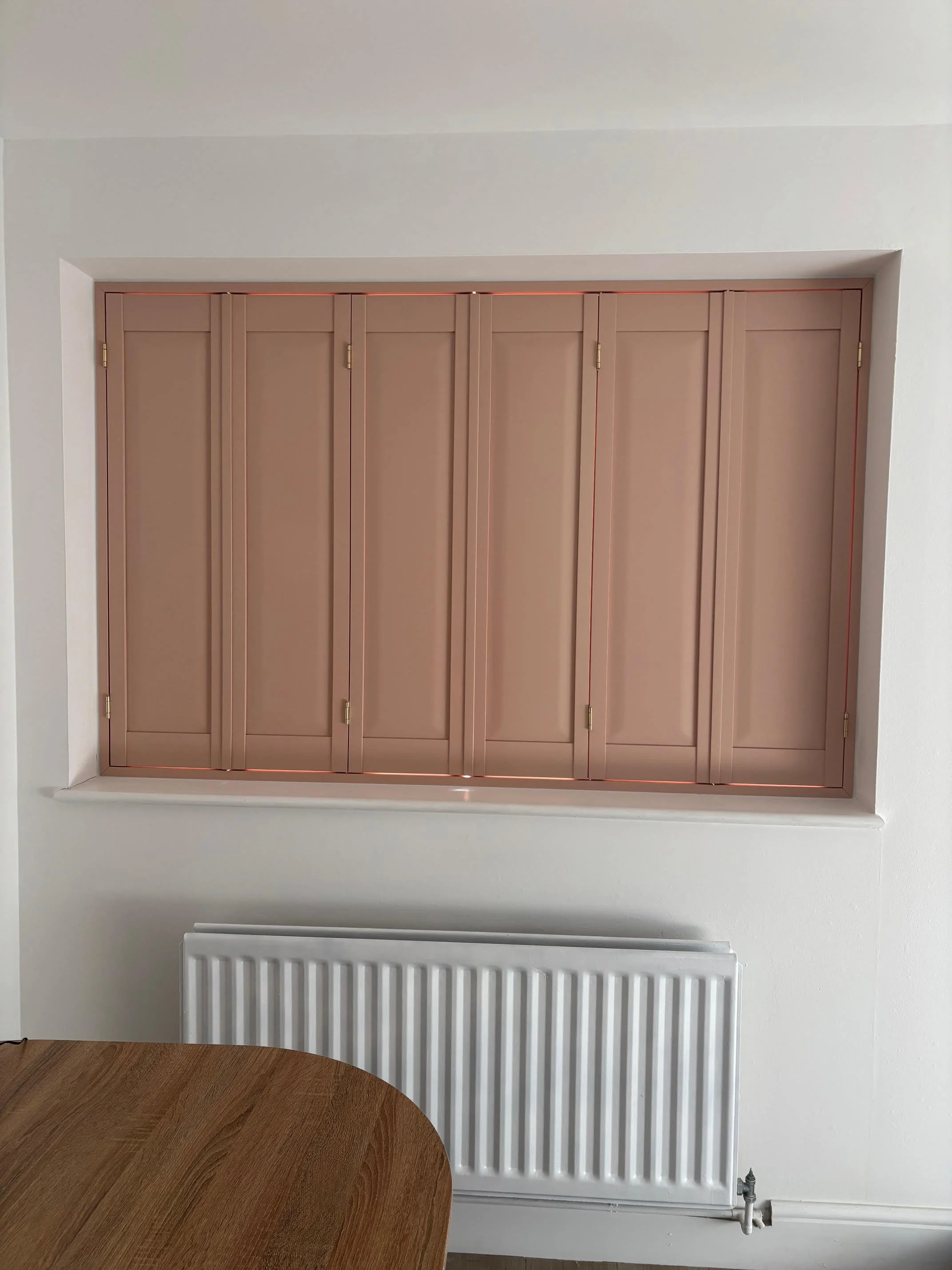A window with closed pink shutters and thin red lines on the edges, positioned above a white radiator and a wooden table in a room with white walls.