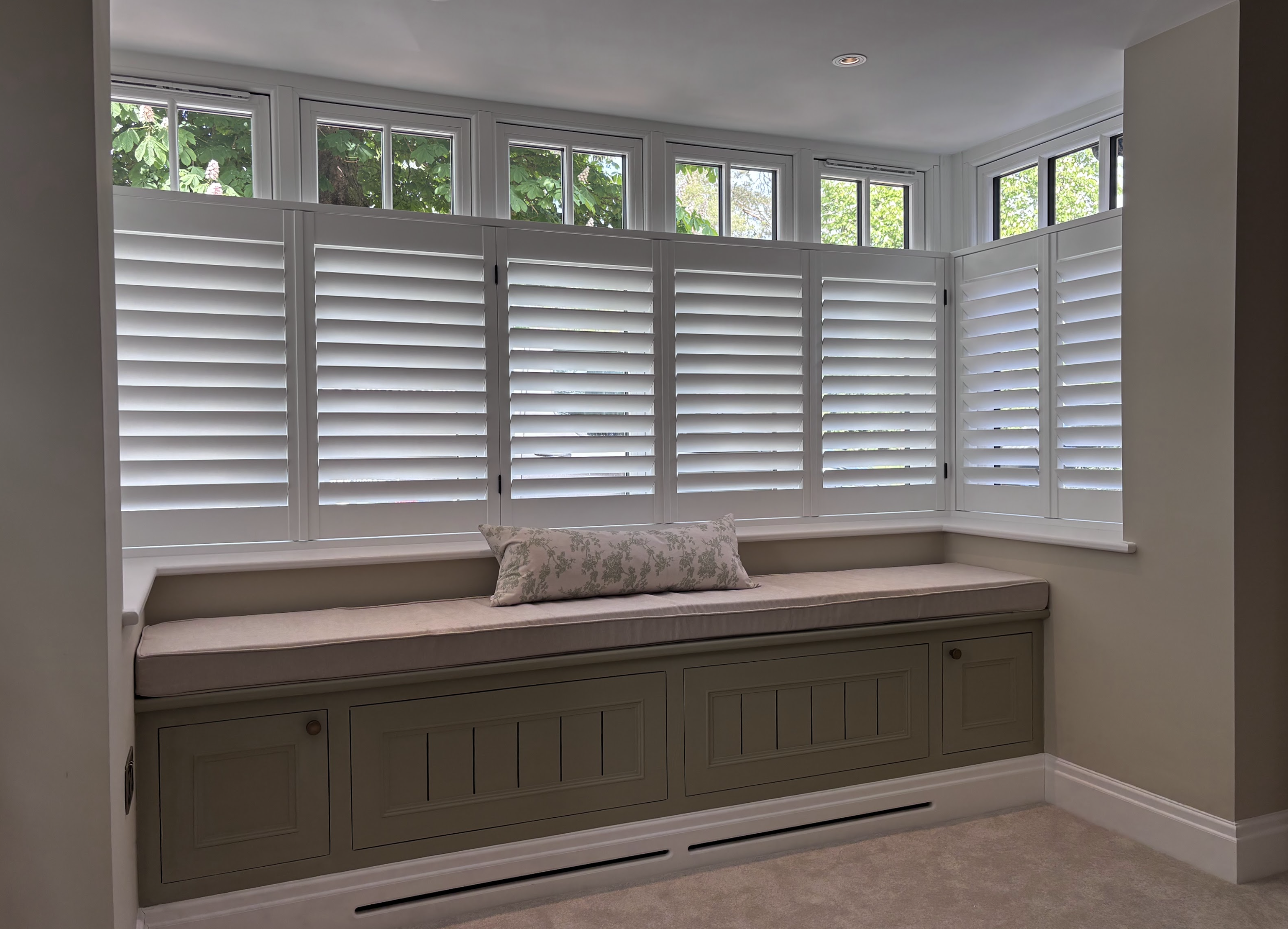 A built-in window seat with white cabinetry and a pink cushion, situated below large corner windows with white plantation shutters, overlooking leafy trees outside.
