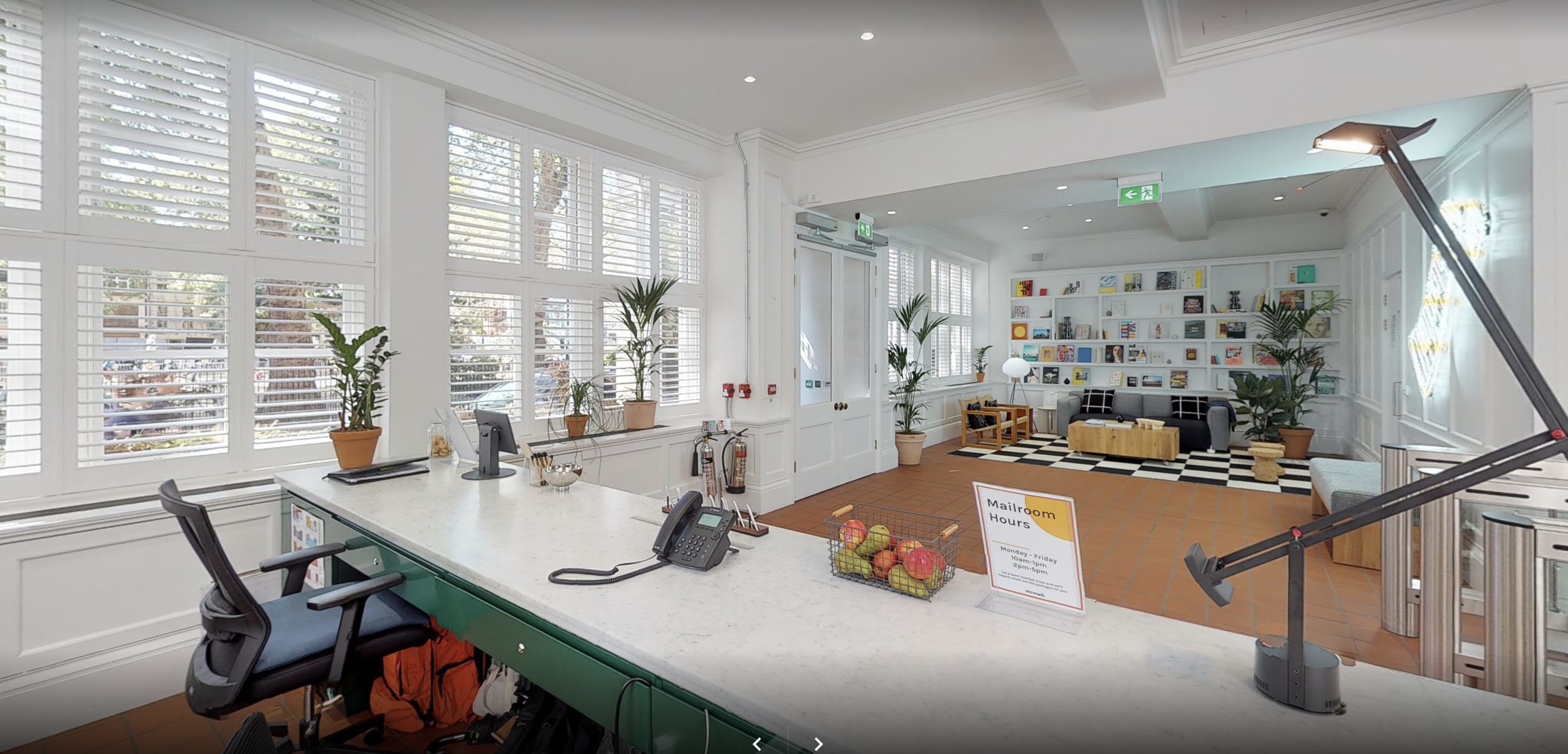 Reception area with large windows, potted plants, a counter with a telephone, a basket of apples, and a sign displaying mailroom hours. In the background, a cozy seating area with a bookshelf, armchair, and checkered rug, illuminated by natural light