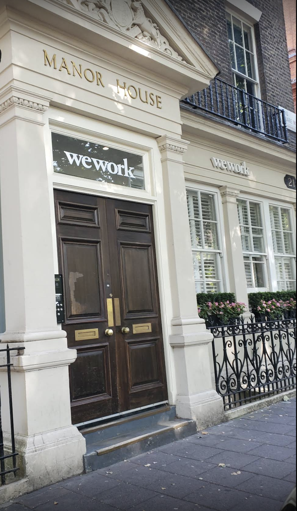 The entrance of a building named Manor House with a dark wooden double door, white trim, and the Wework sign above the door, with a black wrought iron fence in front and flowers in window boxes.