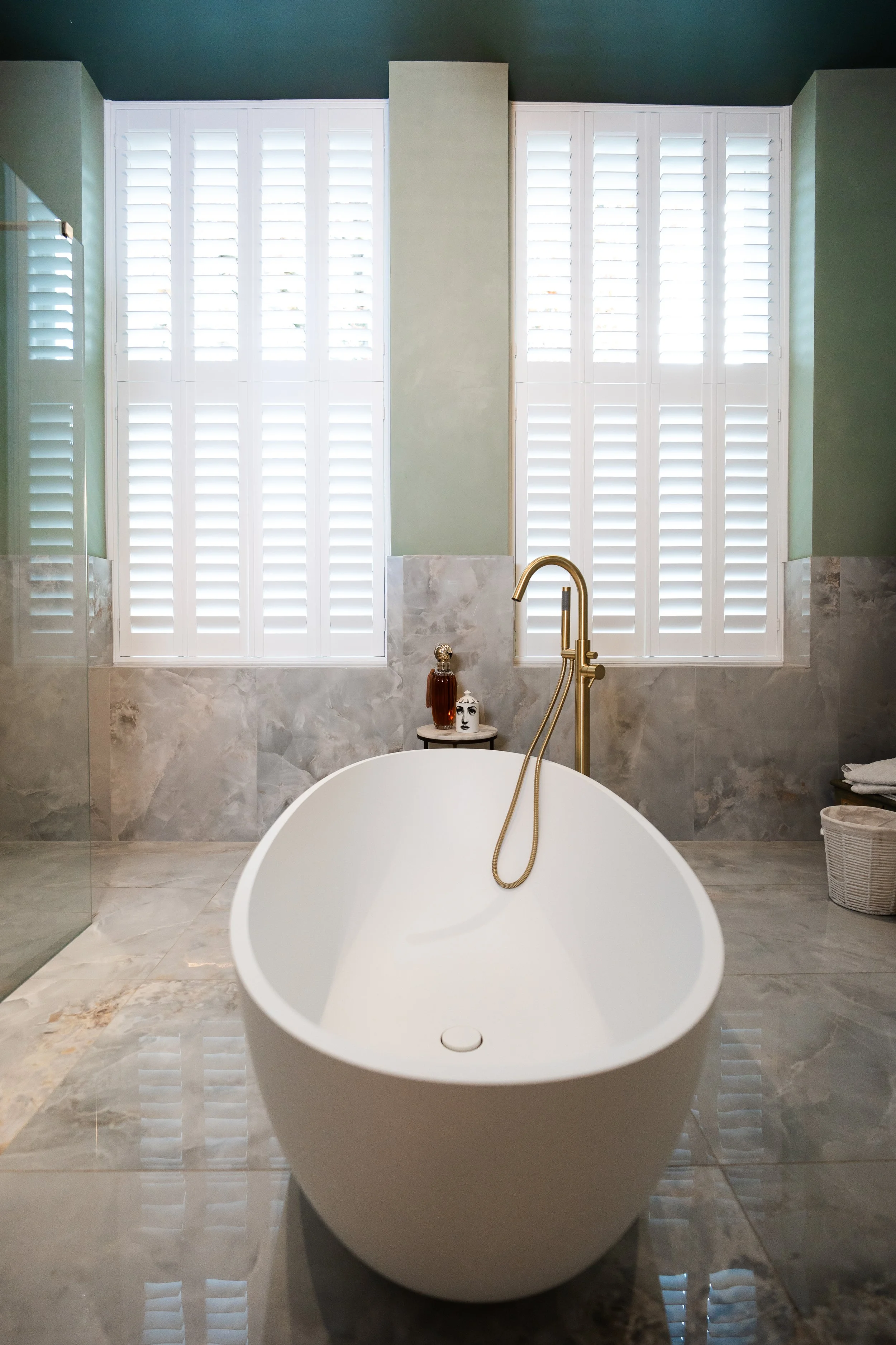 Freestanding bathtub in a modern bathroom with marble tiles, large windows with white shutters, green walls, and brass fixtures.