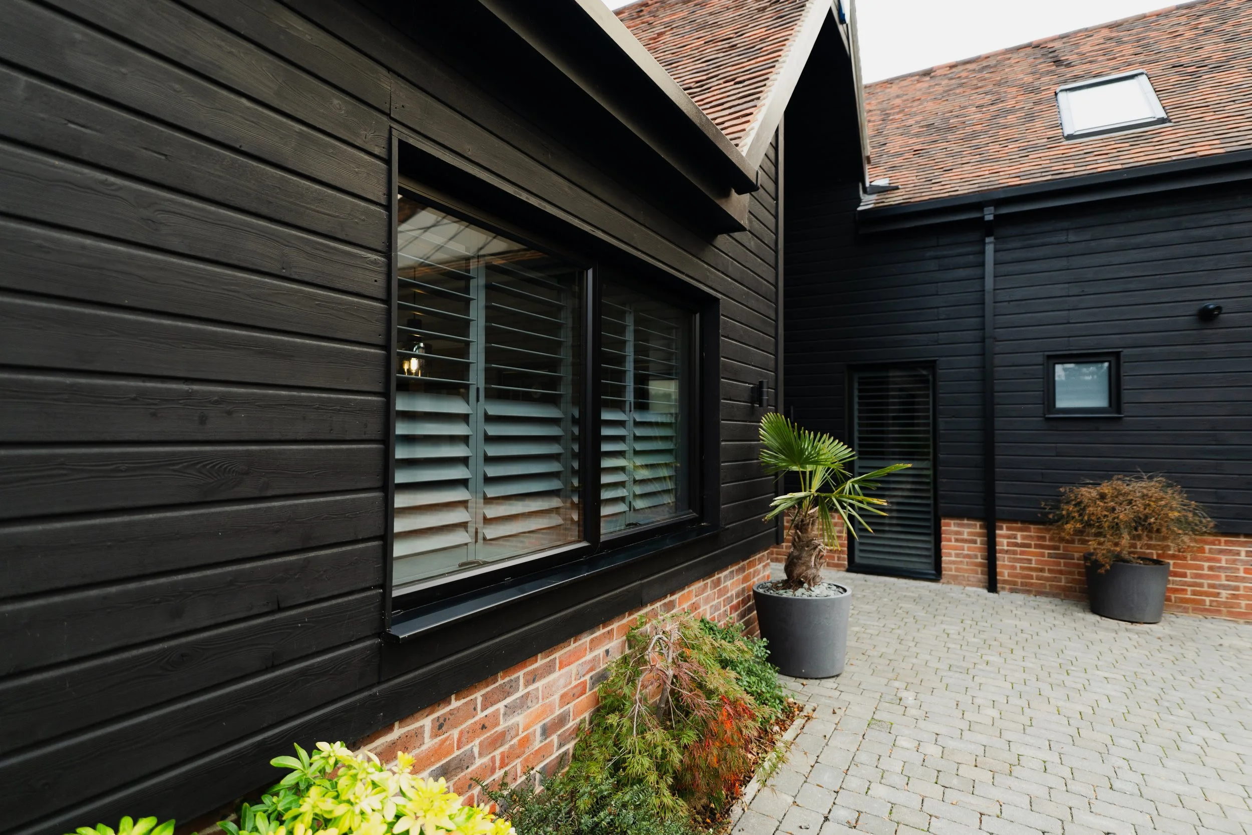 A modern house with dark wooden siding and brick foundation, large window with dark grey window shutters, brick paved walkway, and several potted plants outside.
