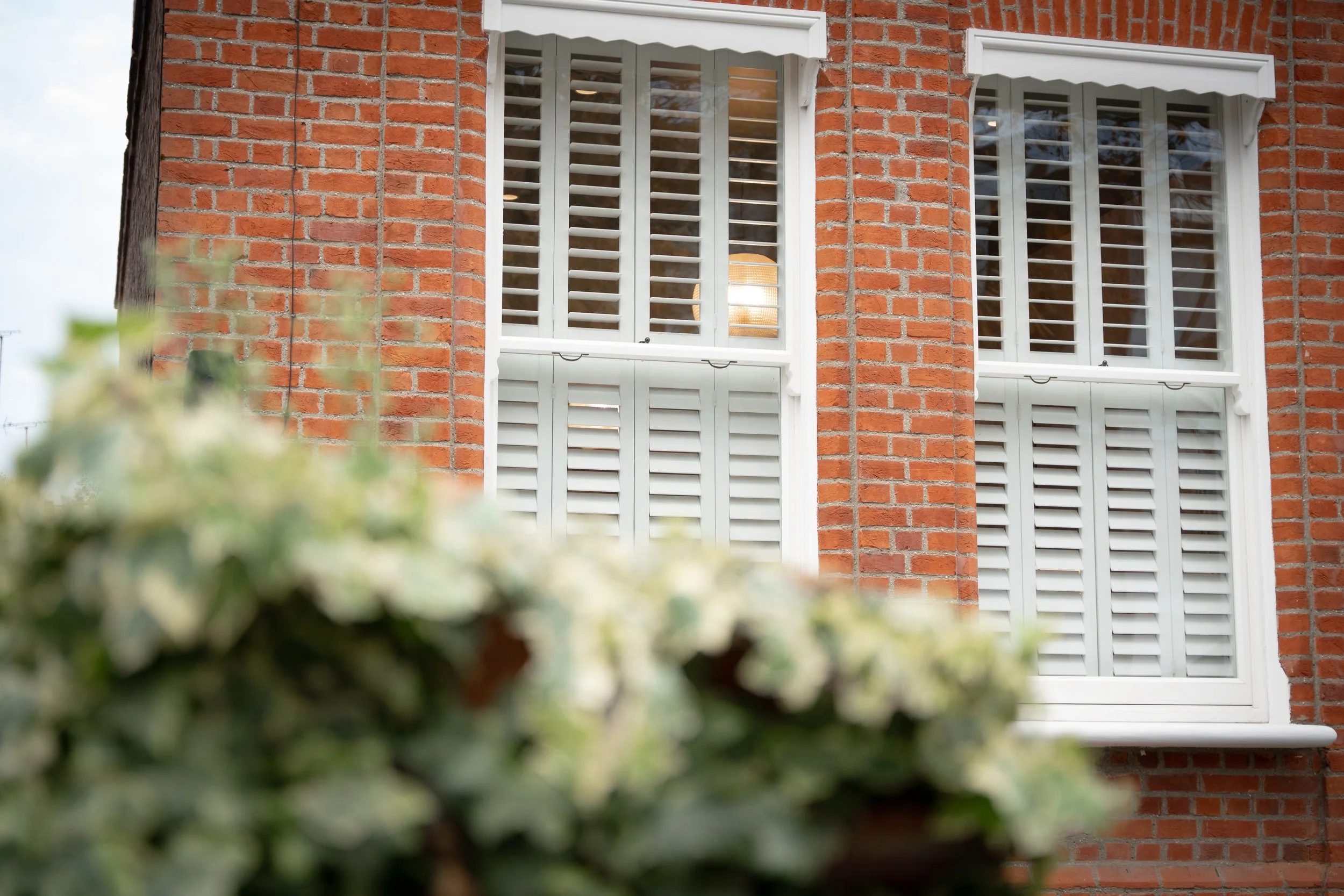 Close-up view of a brick building with two white-framed windows with shutters, viewed through some out-of-focus green foliage in the foreground.