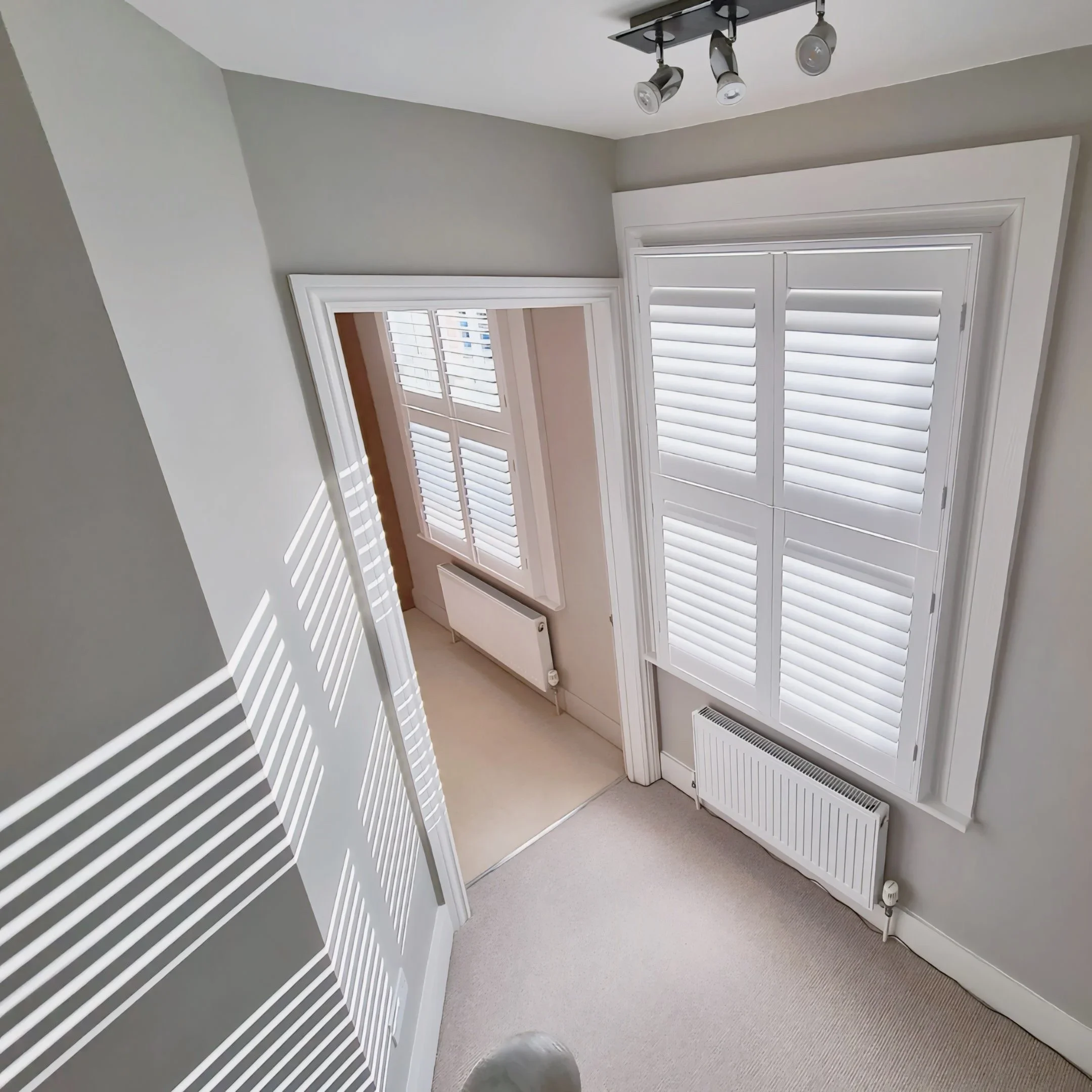 A corner of a room with white walls, white window shutters, a radiator, and a ceiling light fixture with three adjustable spotlights.