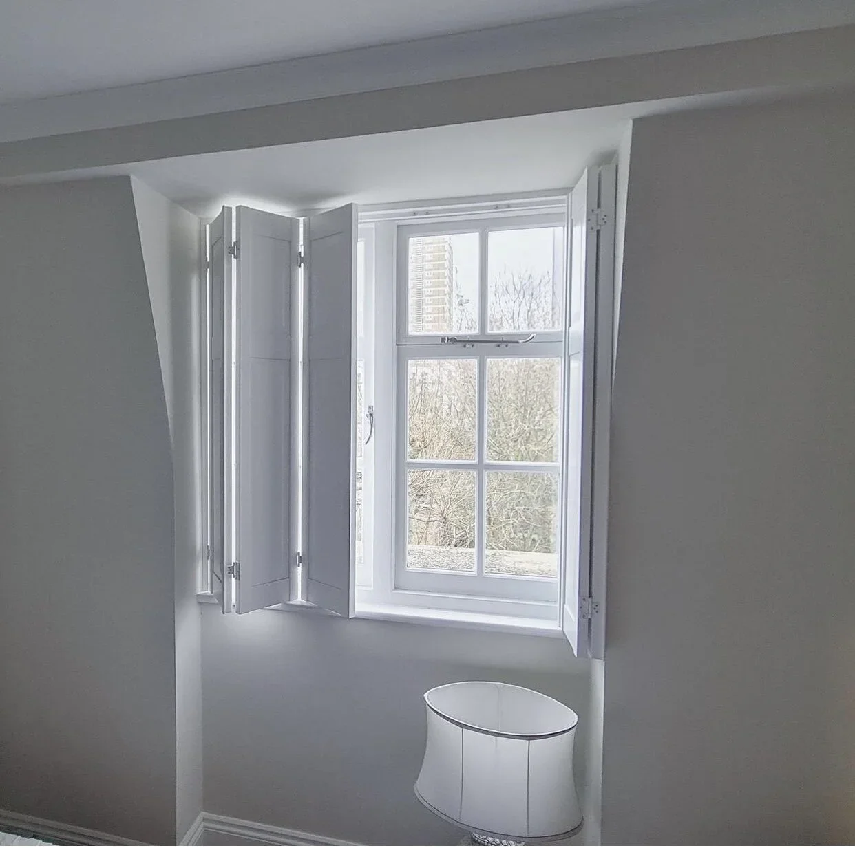 Bright room with a large multi-pane window and white window shutters, showing trees outside on a cloudy day.