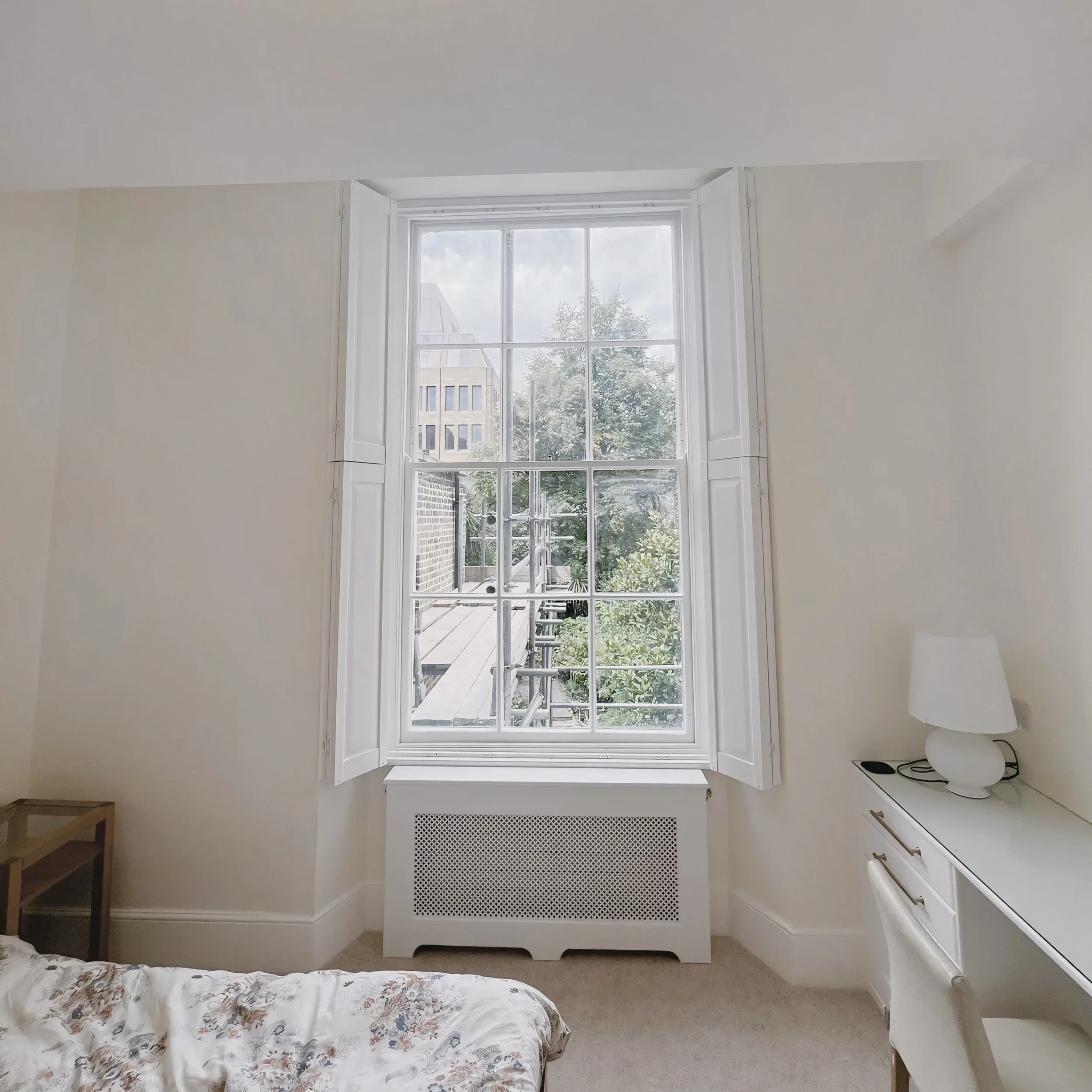 Room with large window, white walls, radiator, small desk, floral bedspread, and a lamp.
