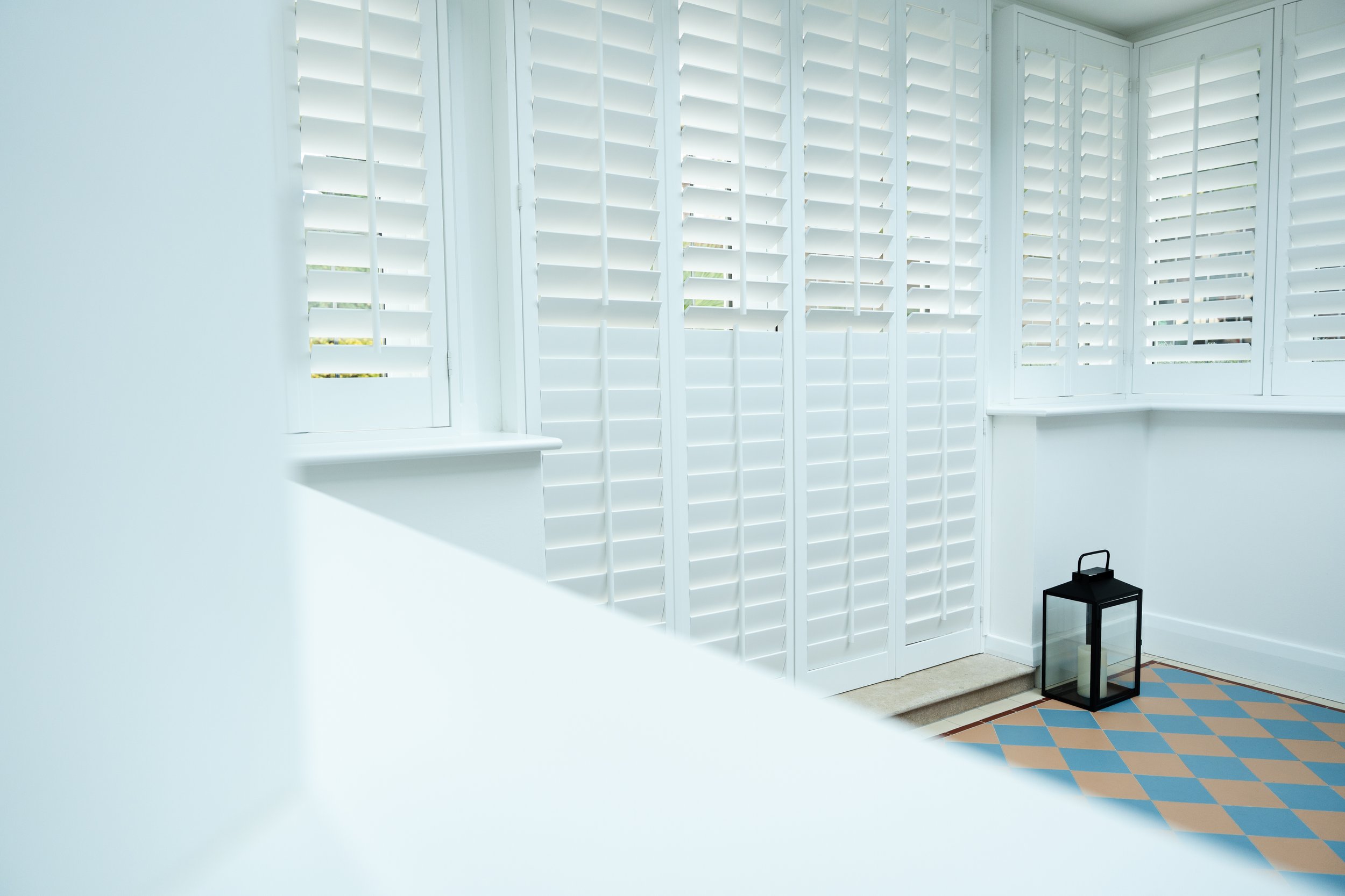 A room with white plantation shutters on the windows, a small black lantern on the floor, and a section of tiled floor with a blue, orange, and beige pattern.