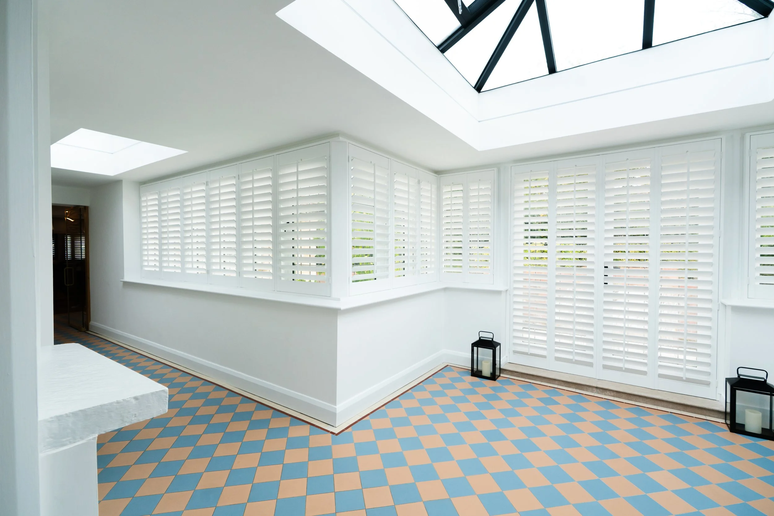 Interior view of a room with white walls and ceiling, large windows with white shutters, a skylight in the ceiling, and a patterned tile floor in shades of blue and beige. There are black lantern-style candle holders with candles on the floor near th