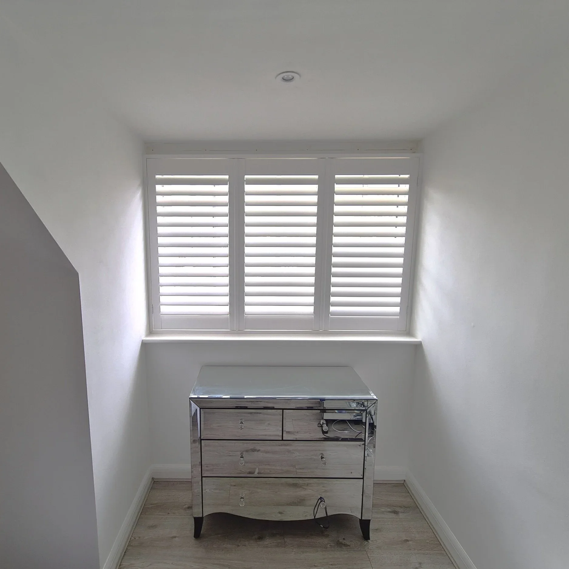 A white room with a window featuring white shutters, a small mirrored dresser with three drawers, and a power strip on the surface. The room has wood-like flooring and white walls.