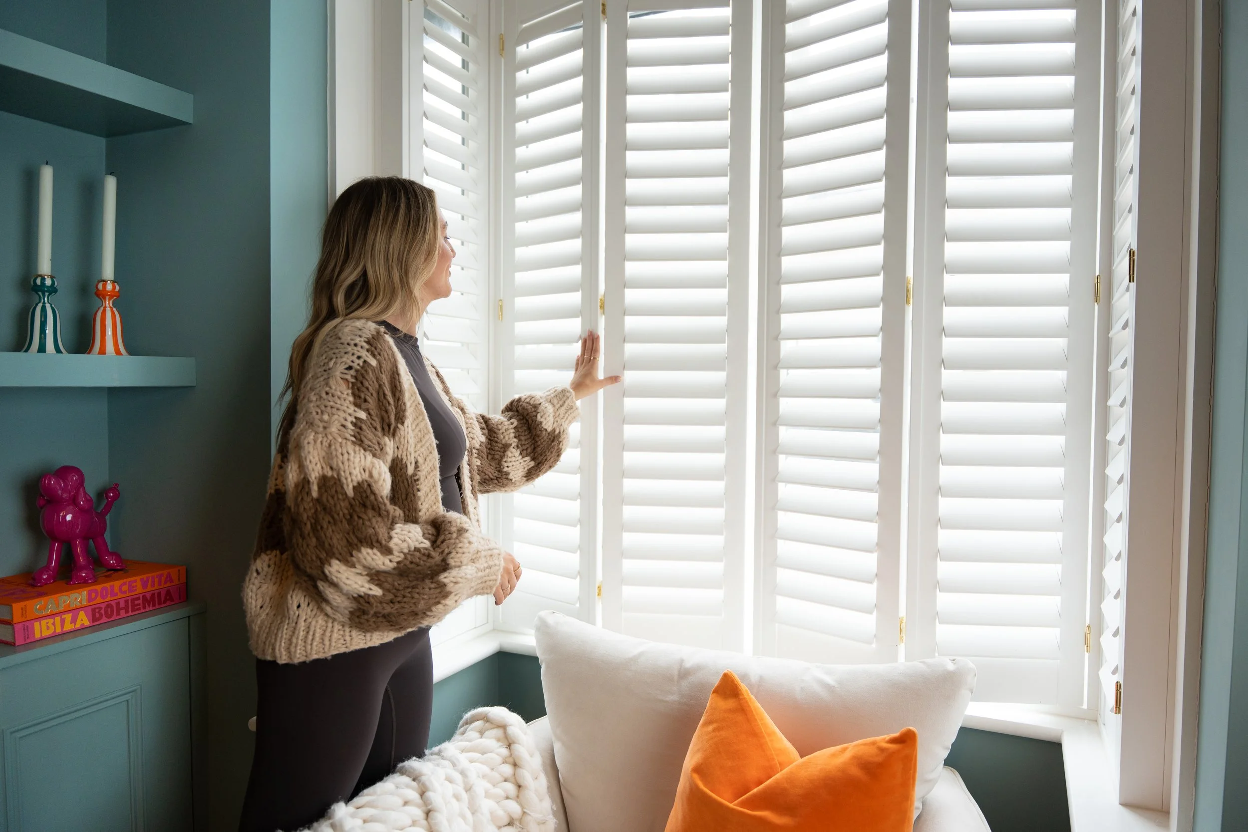 Woman looking out a white window with shutters in a cozy room with teal walls, a forest green shelf holding colorful books and decorative items, a plush white blanket, a beige ottoman, and orange throw pillows.