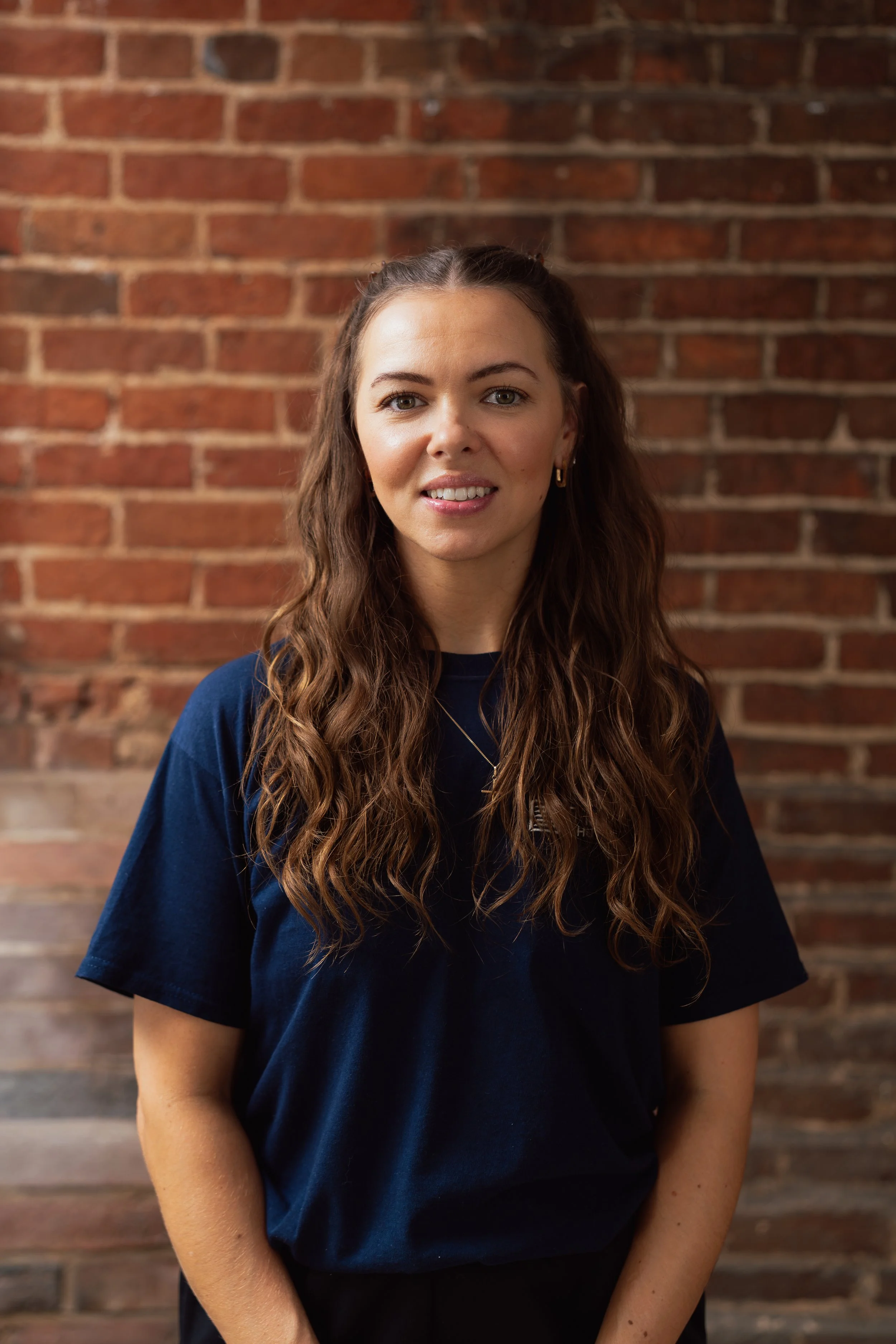 Portrait of a woman with long wavy brown hair, wearing a navy blue T-shirt and gold earrings, standing in front of a brick wall.