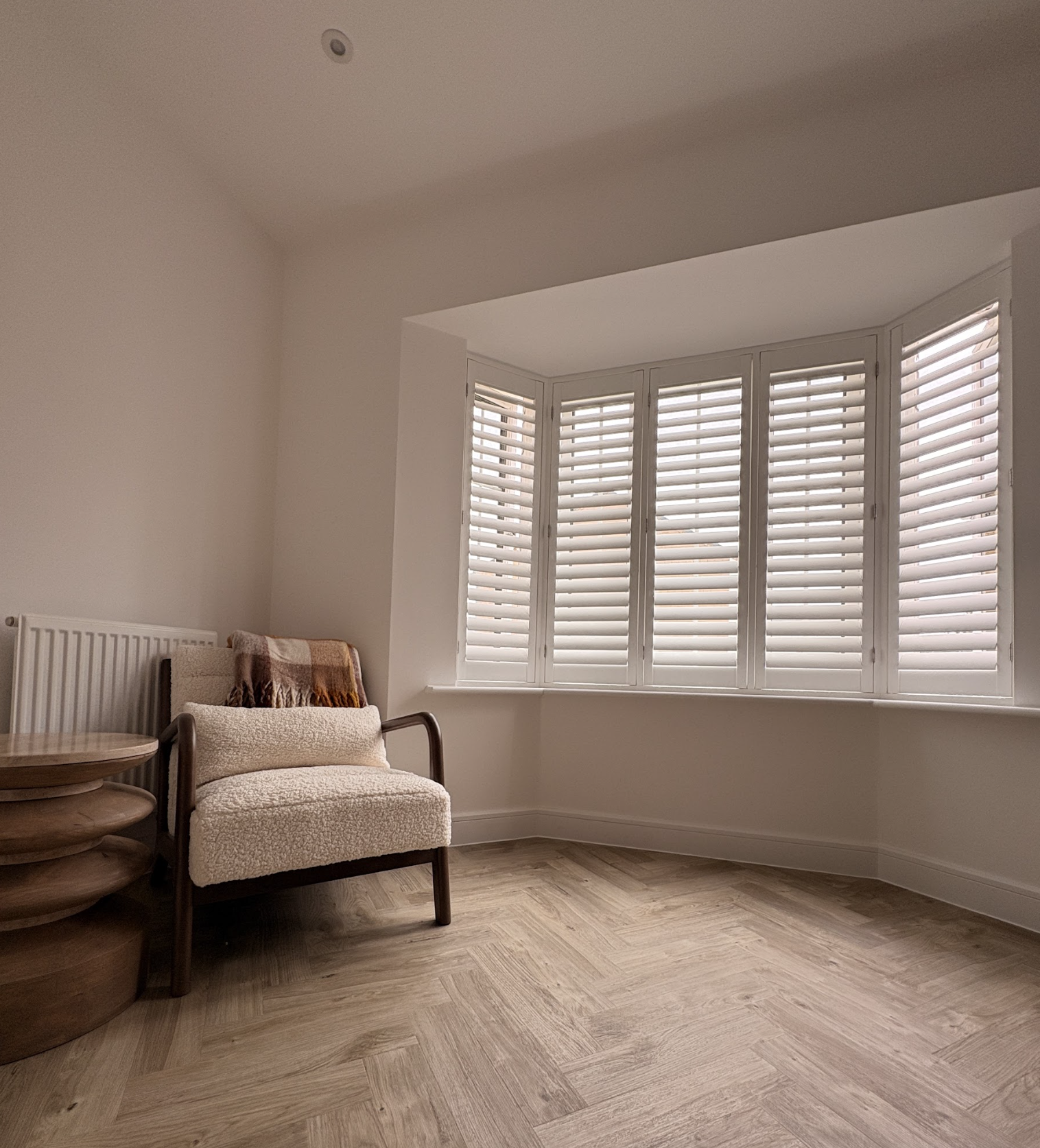 A cozy corner of a living room featuring a cream-colored armchair with a brown checkered blanket draped over the back, next to a small wooden side table, and a large window with white plantation shutters letting in natural light, set on a wood floor.