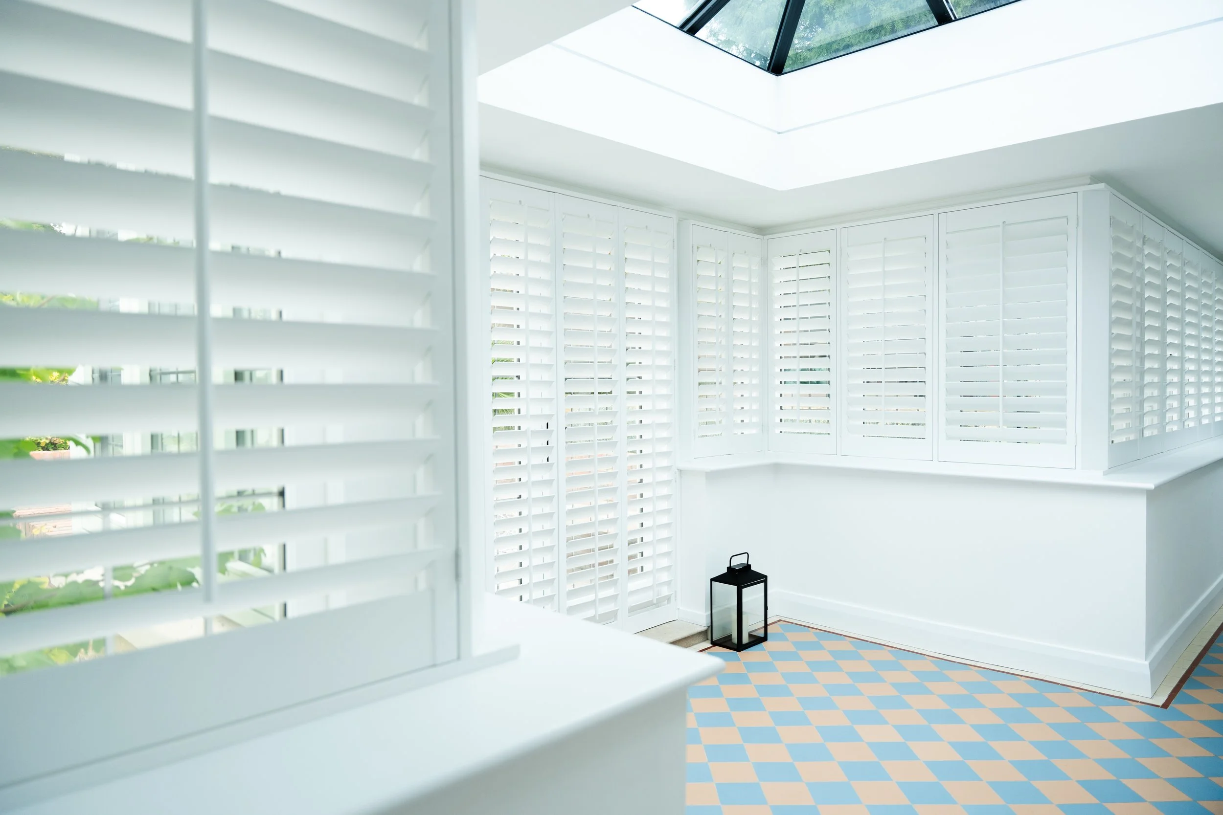 Bright room with white window shutters and a skylight, featuring a corner with a black lantern on the floor and a pastel-colored checkered floor.
