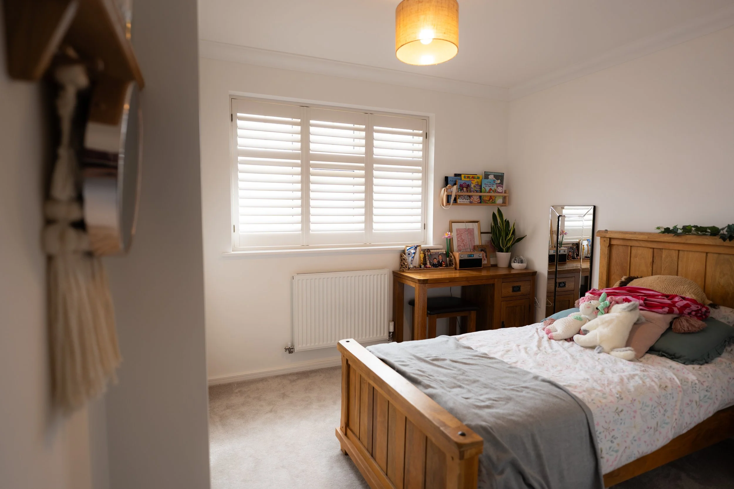 A bedroom with a large window with white shutters, a wooden bed frame with a floral bedspread and stuffed animals, a wooden desk with framed photographs, a potted plant, and a small mirror. A ceiling light fixture illuminates the room.