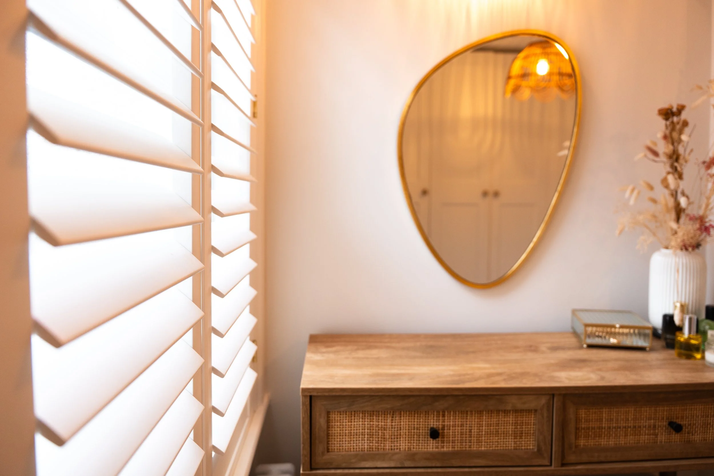 A mirror with a gold frame hangs on a white wall in a room with beige walls. There is a wooden dresser below the mirror, and a white vase containing dried flowers is placed on top. To the left, white plantation shutters cover a window, allowing some 
