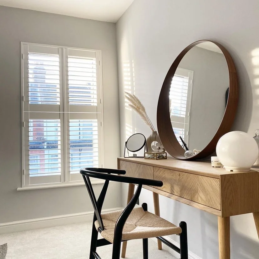 A minimalist bedroom or dressing area with a light wood vanity or desk, decorated with a large circular mirror, a small round mirror, a white ceramic vase with pampas grass, and other decorative items. A black chair with a woven seat is in front of the desk, and window shutters allow natural light into the room.