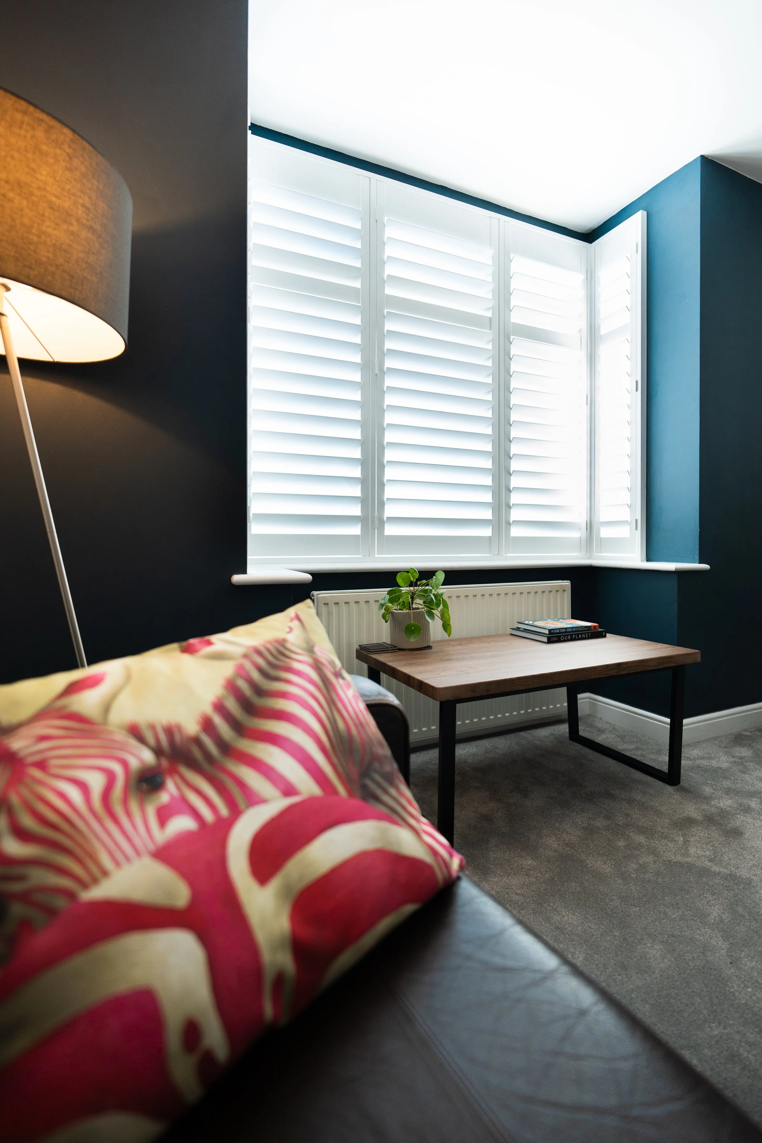 Living room with dark blue wall, large window with white shutters, bed with pink and cream patterned pillow, small wooden table with a potted plant, books, and electronic device, gray carpet, and a white radiator.