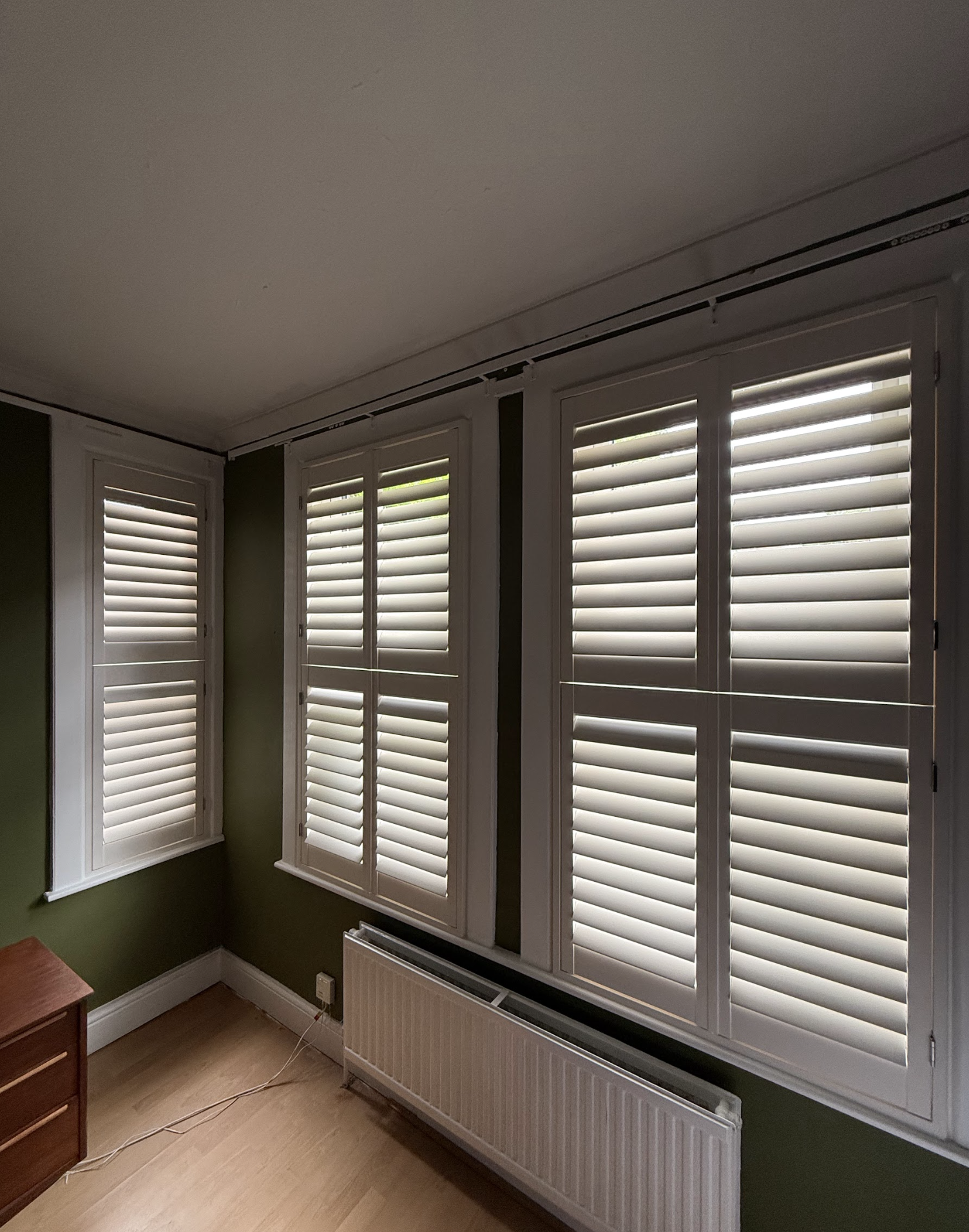 Room with three large windows fitted with white horizontal blinds, green walls, a radiator beneath the windows, and a wooden dresser on the left side.