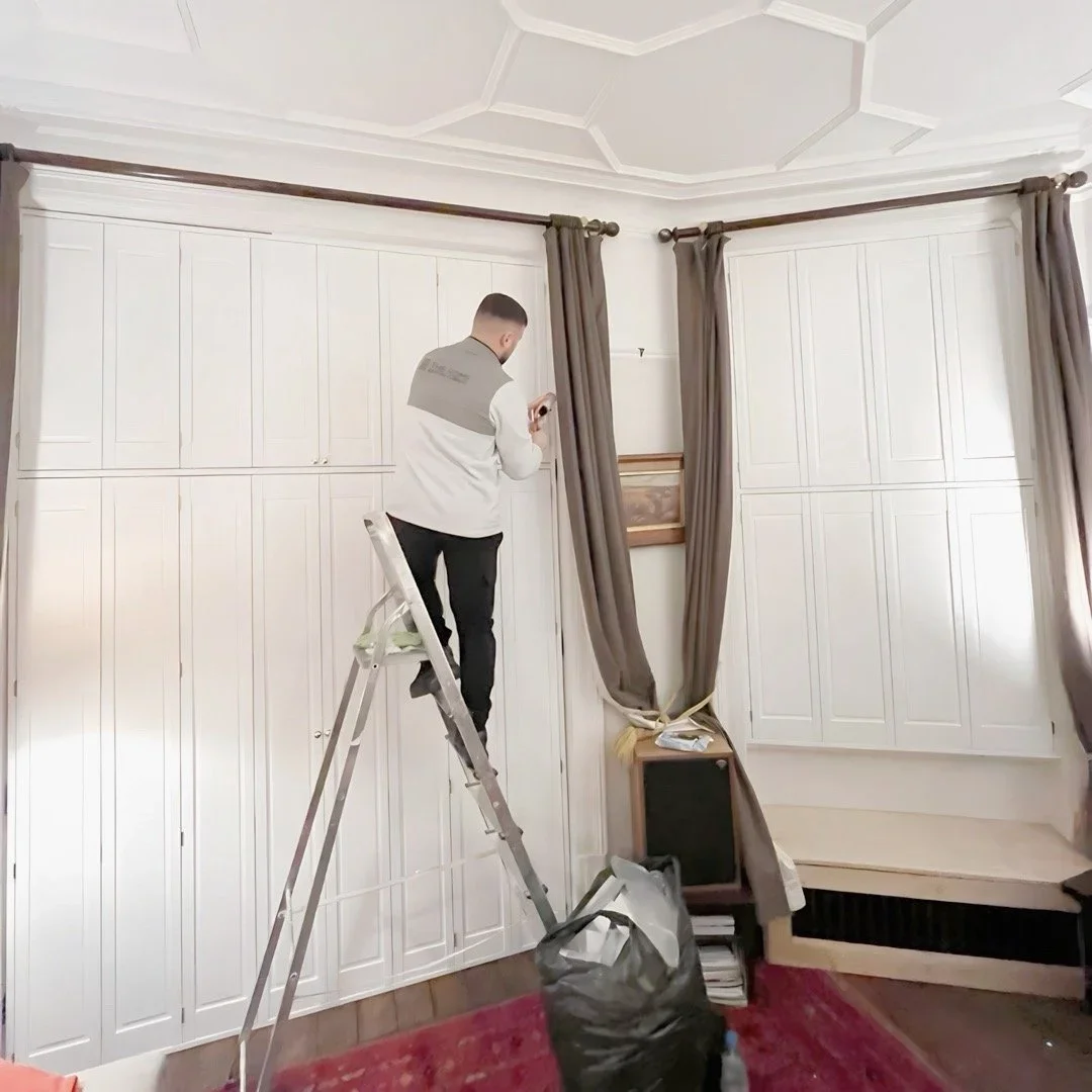 A man standing on a ladder installing or adjusting shutters in a room with white built-in cabinets and a small fireplace.