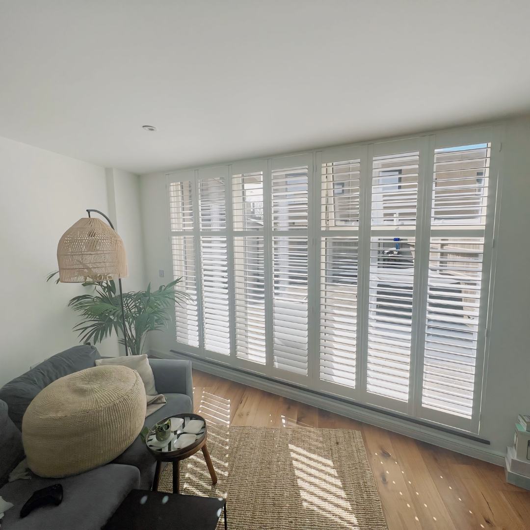 Bright living room with large windows, white shutters, gray sofa with pillows, round wicker ottoman, floor lamp with woven shade, wooden flooring, and a woven rug.