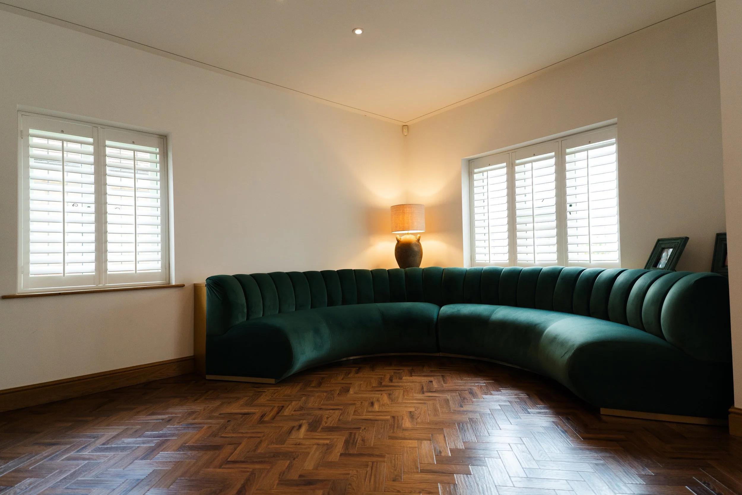 Living room with two windows with white shutters, a green velvet curved sofa, a wooden side table with a lamp, framed photos, and wooden parquet flooring.