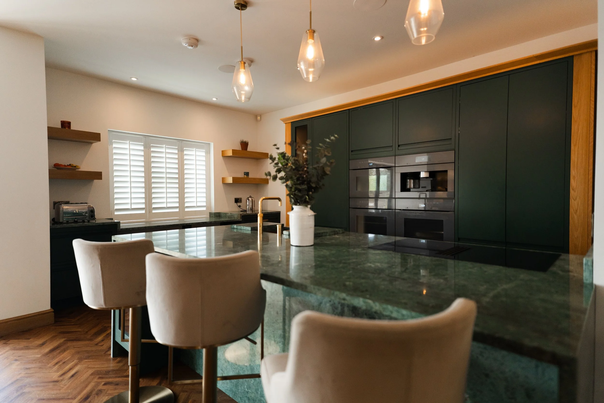Modern kitchen with green cabinets, a black countertop, and a dining table with beige chairs. Pendant lights hang from the ceiling, and decorative shelves are mounted on the wall. Window with white shutters lets in natural light.