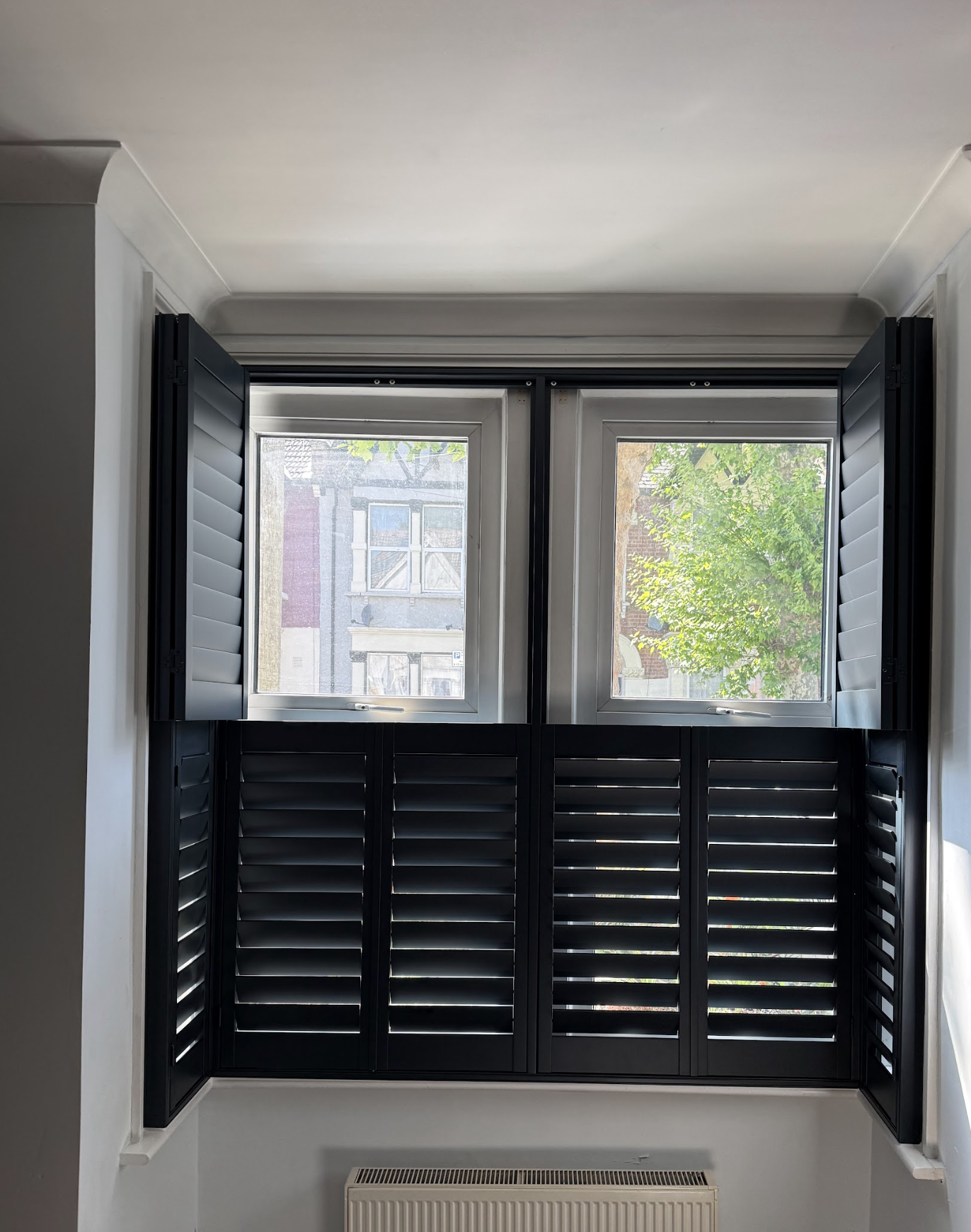 A window with black shutters opened outward, revealing a view of a building and green trees outside, with sunlight illuminating the interior of the room.