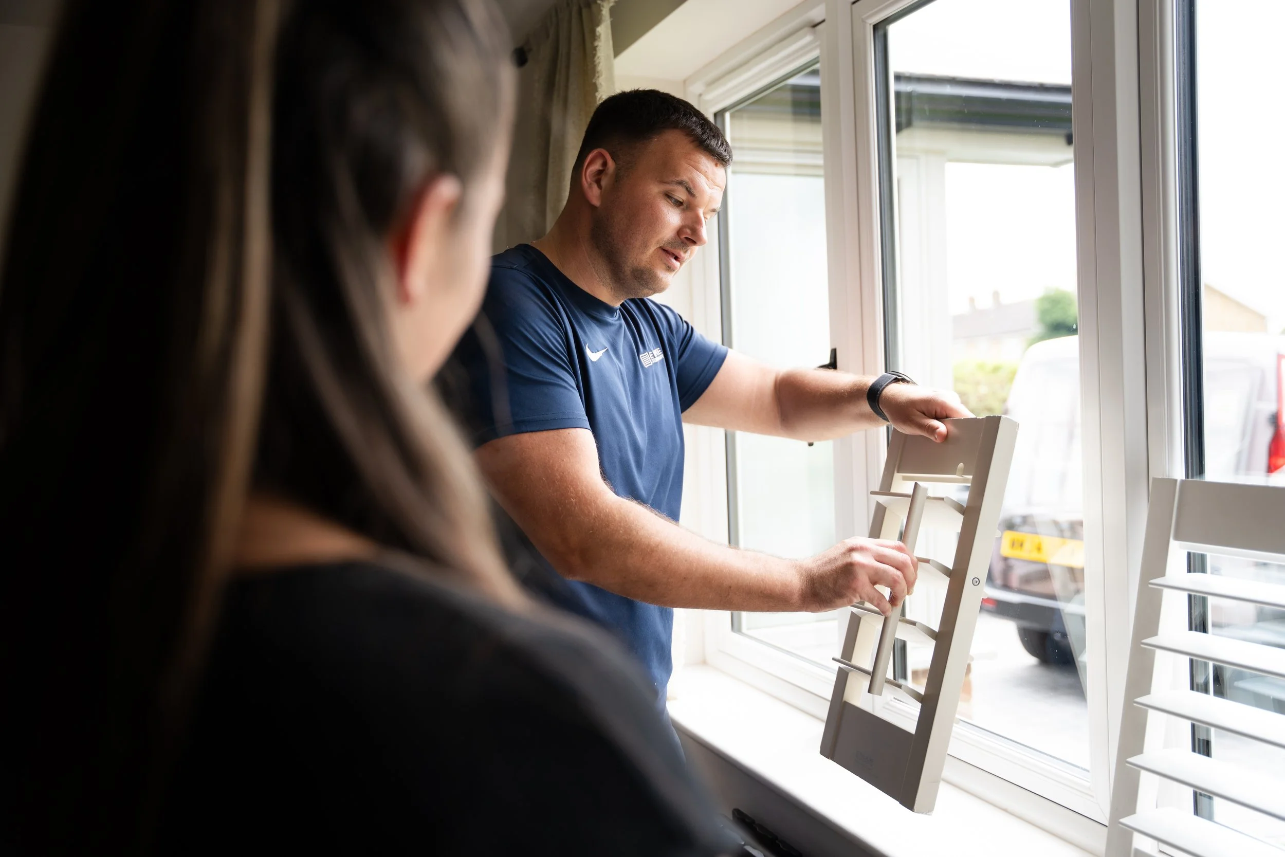 A man wearing a navy blue shirt adjusting a white window blind inside a room, with a view of the outside through the window.