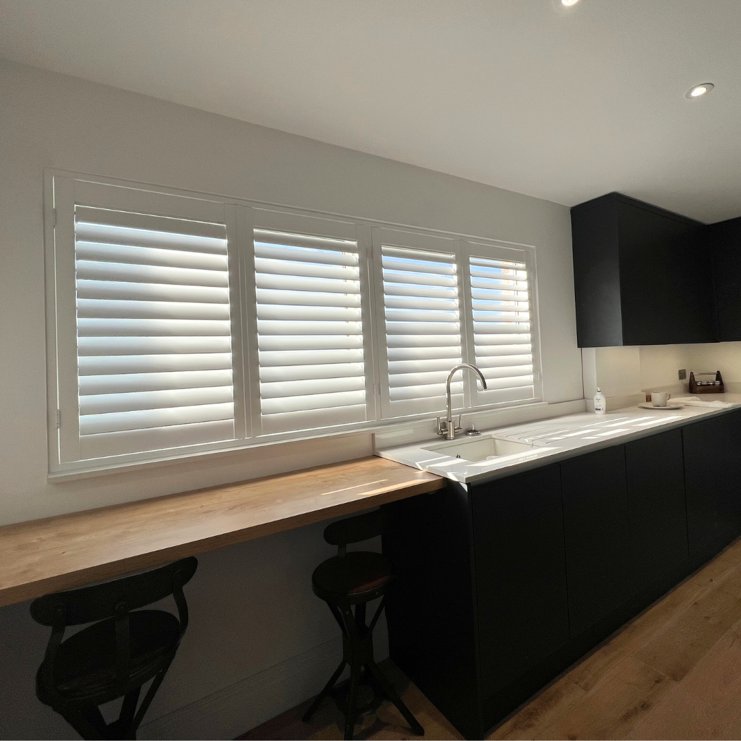 Modern kitchen with white walls, a row of large window blinds, black cabinets, a white countertop, a stainless steel sink with a faucet, and two black barstools along a wooden breakfast bar.