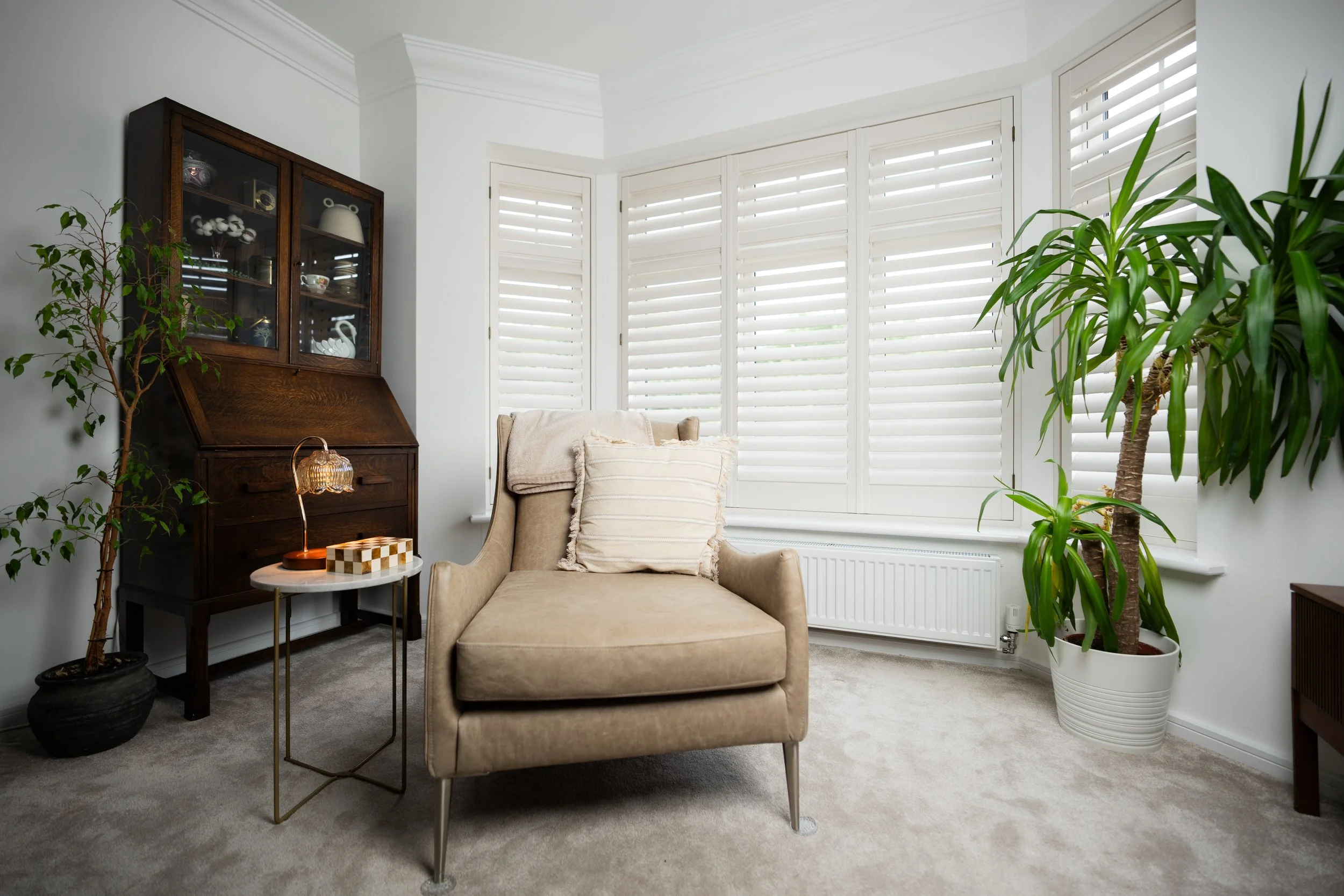 Living room with beige armchair, side table with a lamp, potted plants, a wooden cabinet, and large windows with white shutters.