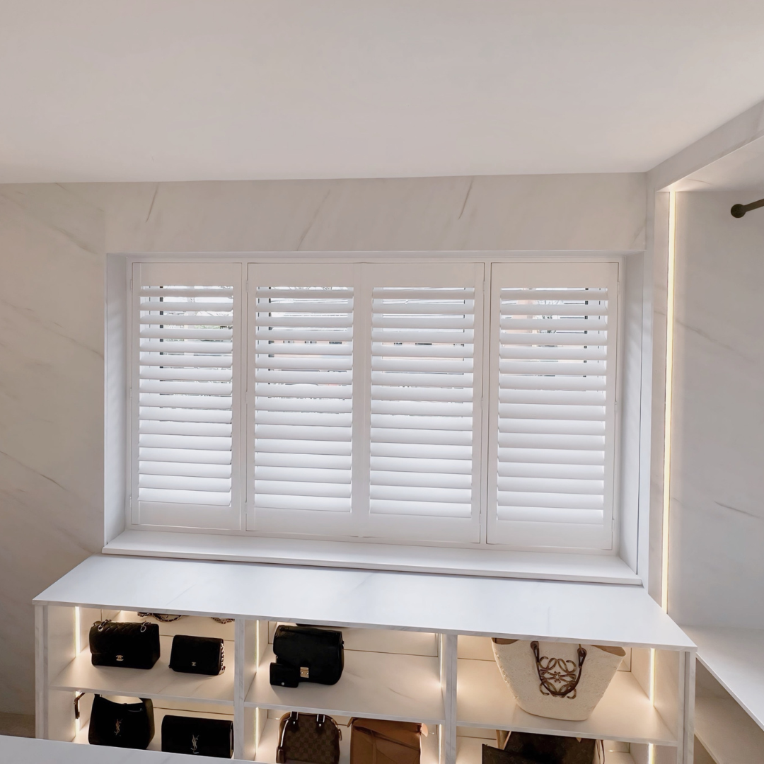 White plantation shutters covering a large window with a surrounding white marble frame. Below, a white shelf displays handbags, some with illuminated backing.