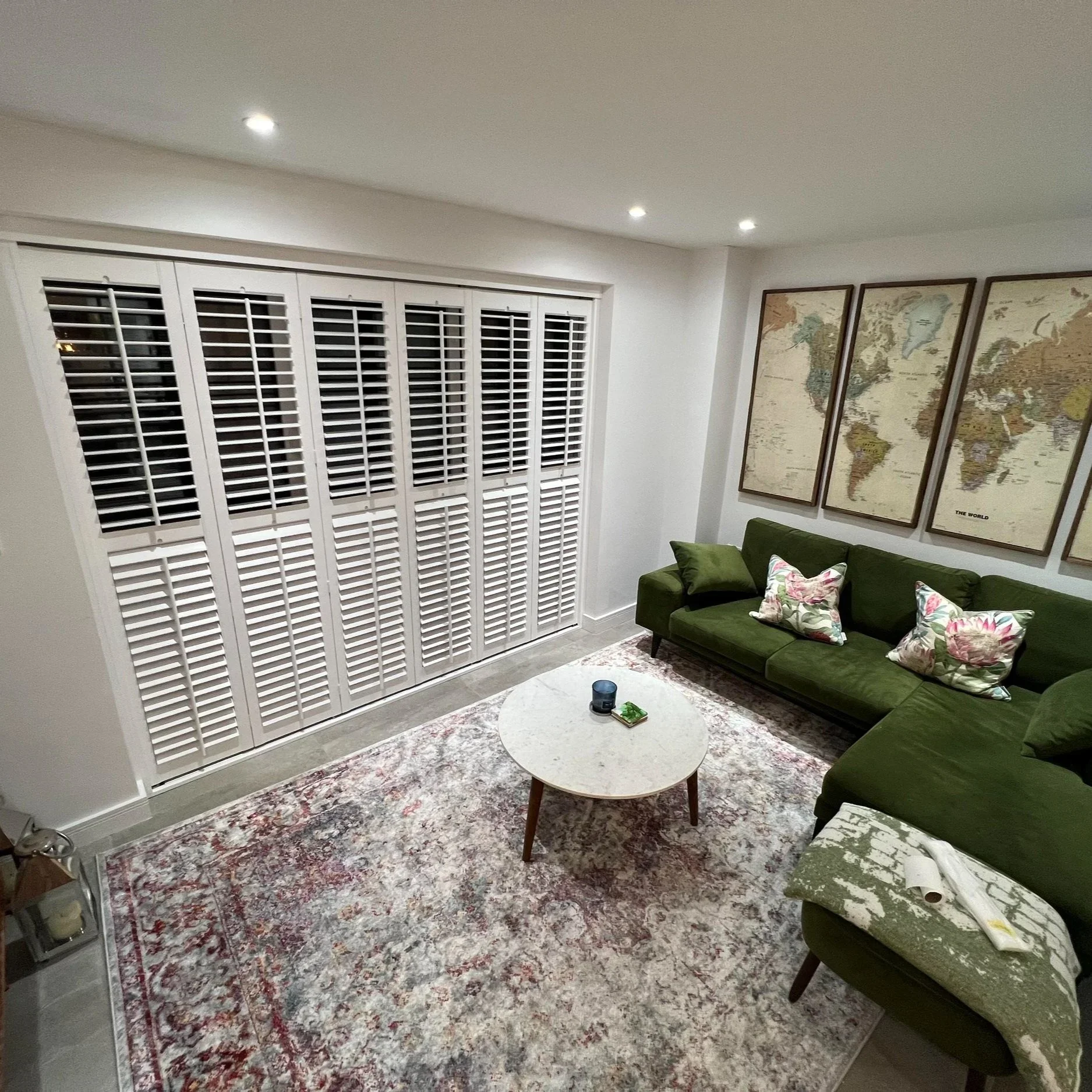 Living room interior with a white window with shutters, framed world maps on the wall, a dark green sofa with floral cushions, a small round white coffee table, a colorful area rug, and ceiling lights.