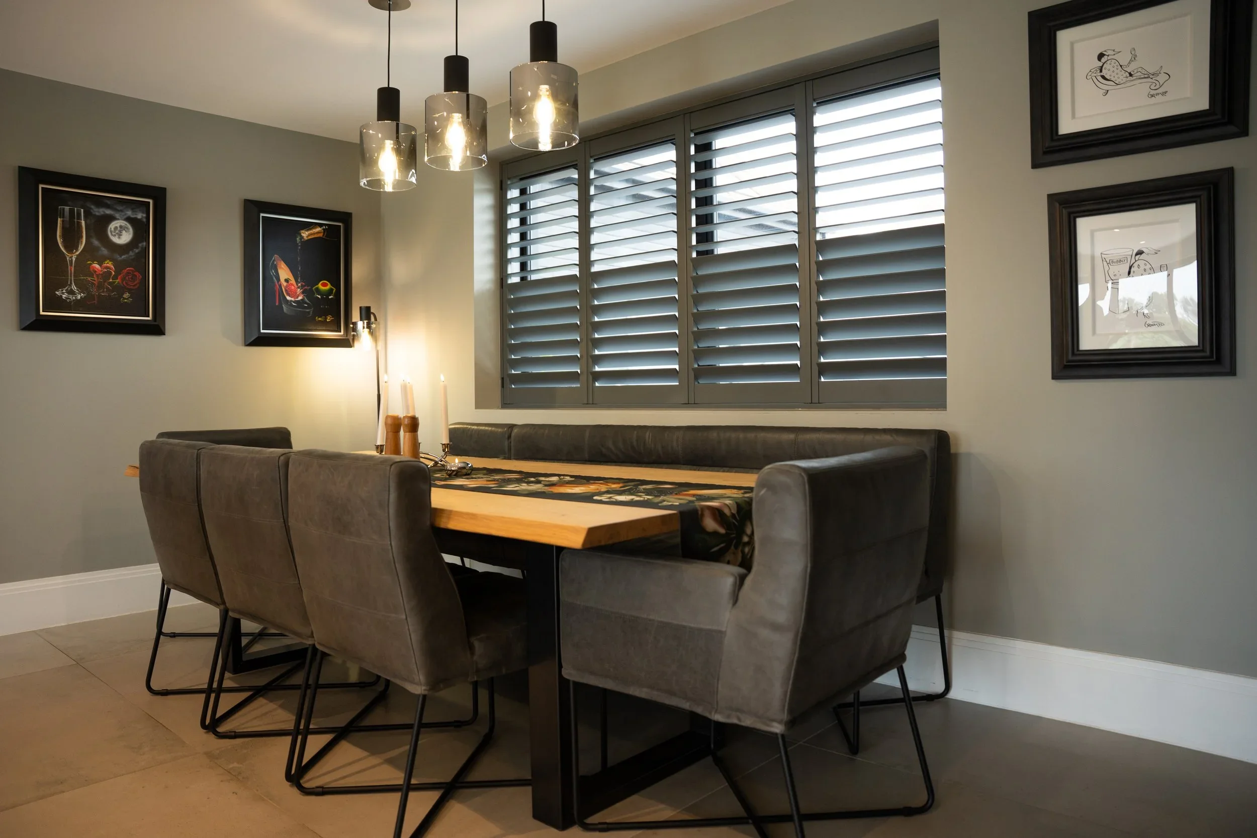 Dining room with a wooden table surrounded by gray upholstered chairs, wall art, and a window with gray shutters.