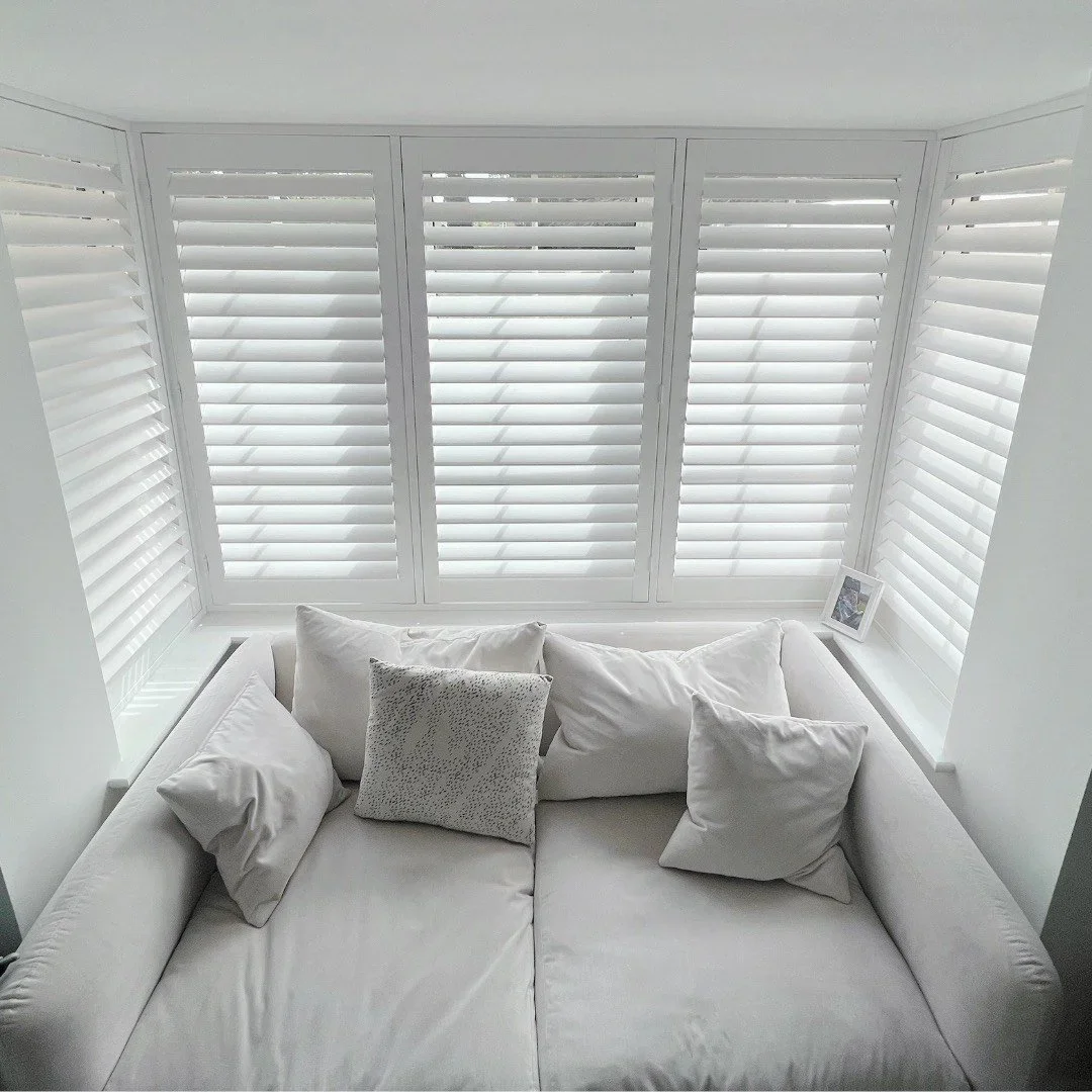 White sofa with multiple white and gray pillows in front of large windows with white plantation shutters.