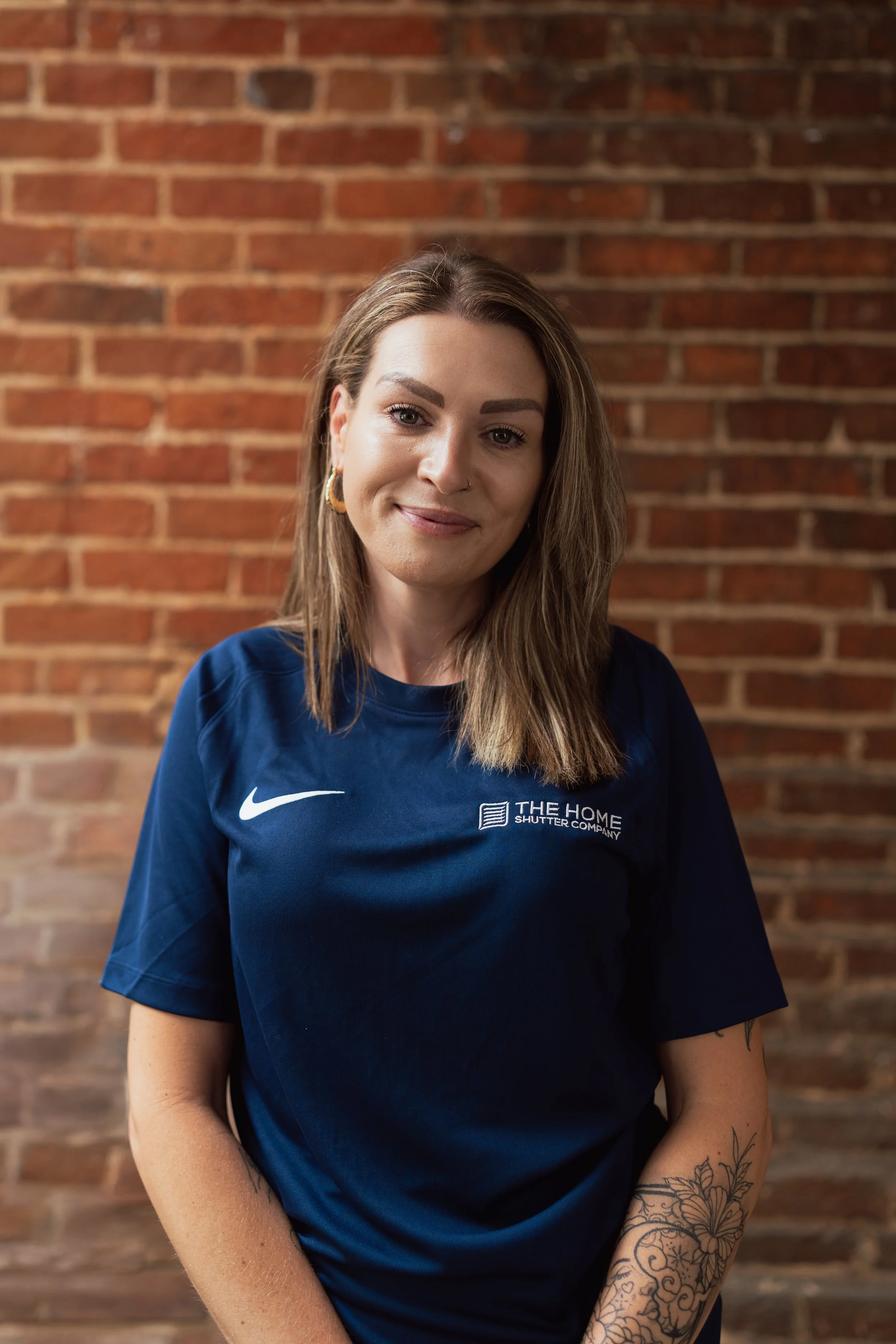 A woman with long brown hair, wearing a blue shirt with logos, standing against a brick wall. She has earrings, a nose ring, and tattoos on her left arm.