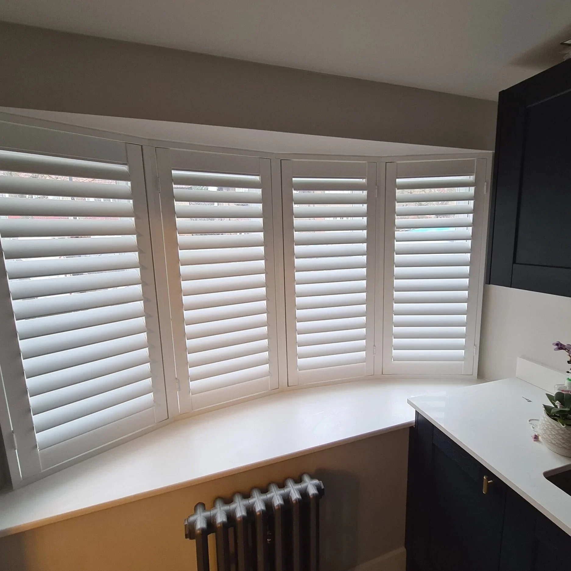 A window with white plantation shutters, a wide window sill, and a view outside, with part of a radiator and a black cabinet nearby